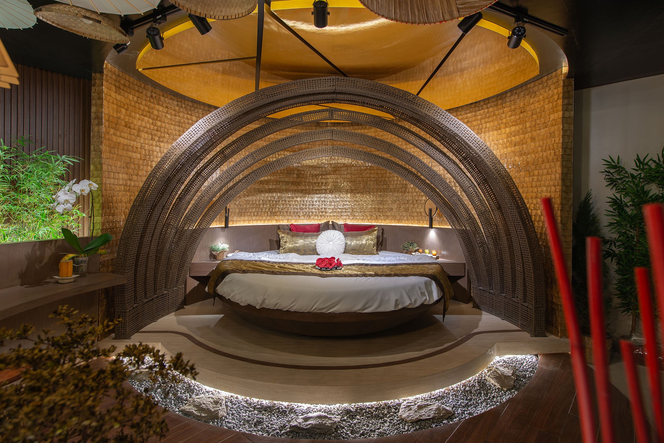 A luxurious bed with white sheets, topped with gold and velvet pillows and a white round pillow in a modern bedroom with a circular design. The bed is framed by a large, semi-circular woven structure. The room features warm yellow lighting, textured gold walls, and surrounding green plants, creating a cozy and elegant ambiance.