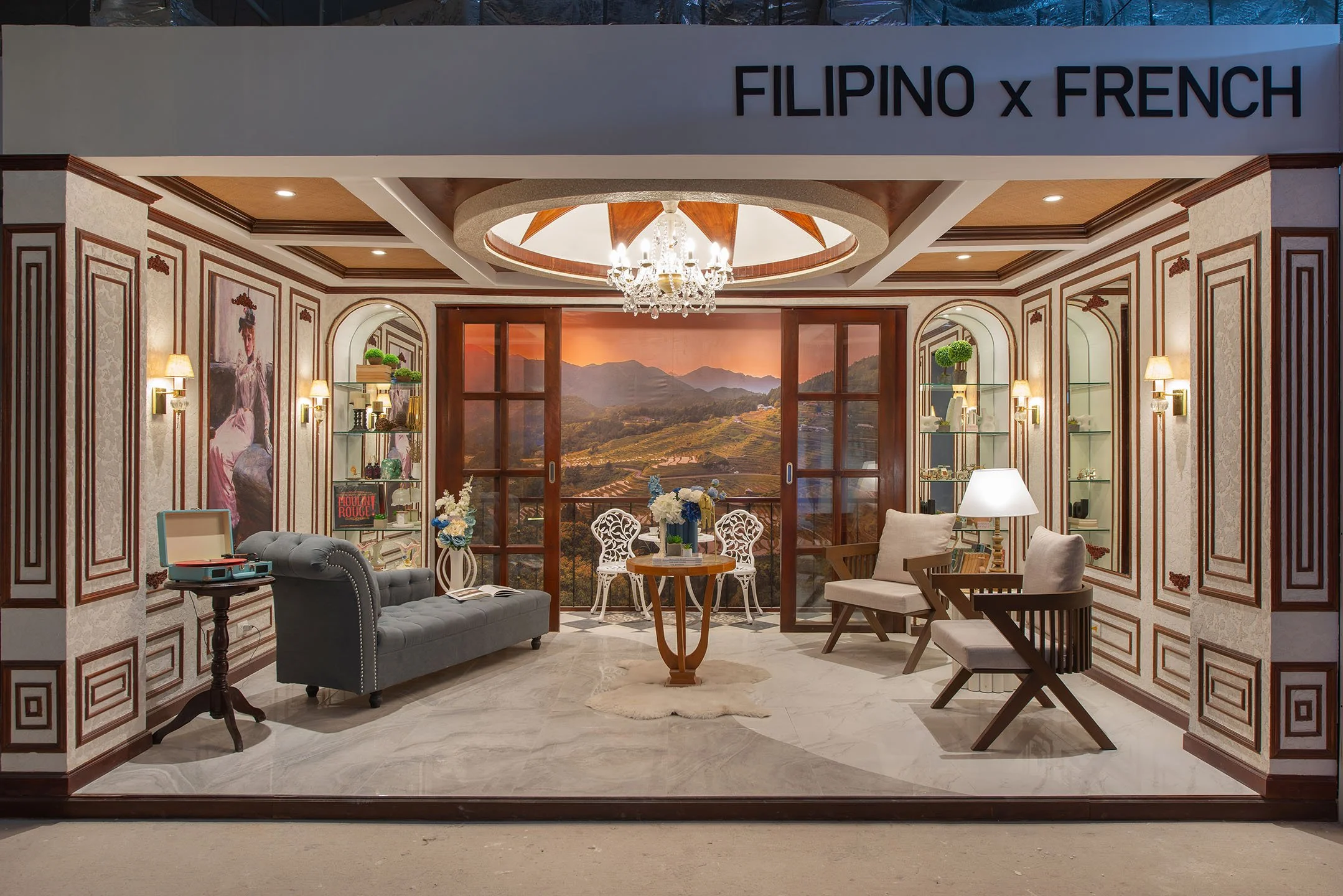 Interior of a room with vintage and modern decor, featuring a chandelier, framed artwork, shelves with decorative items, and a large window showing a scenic mountain view at sunset.