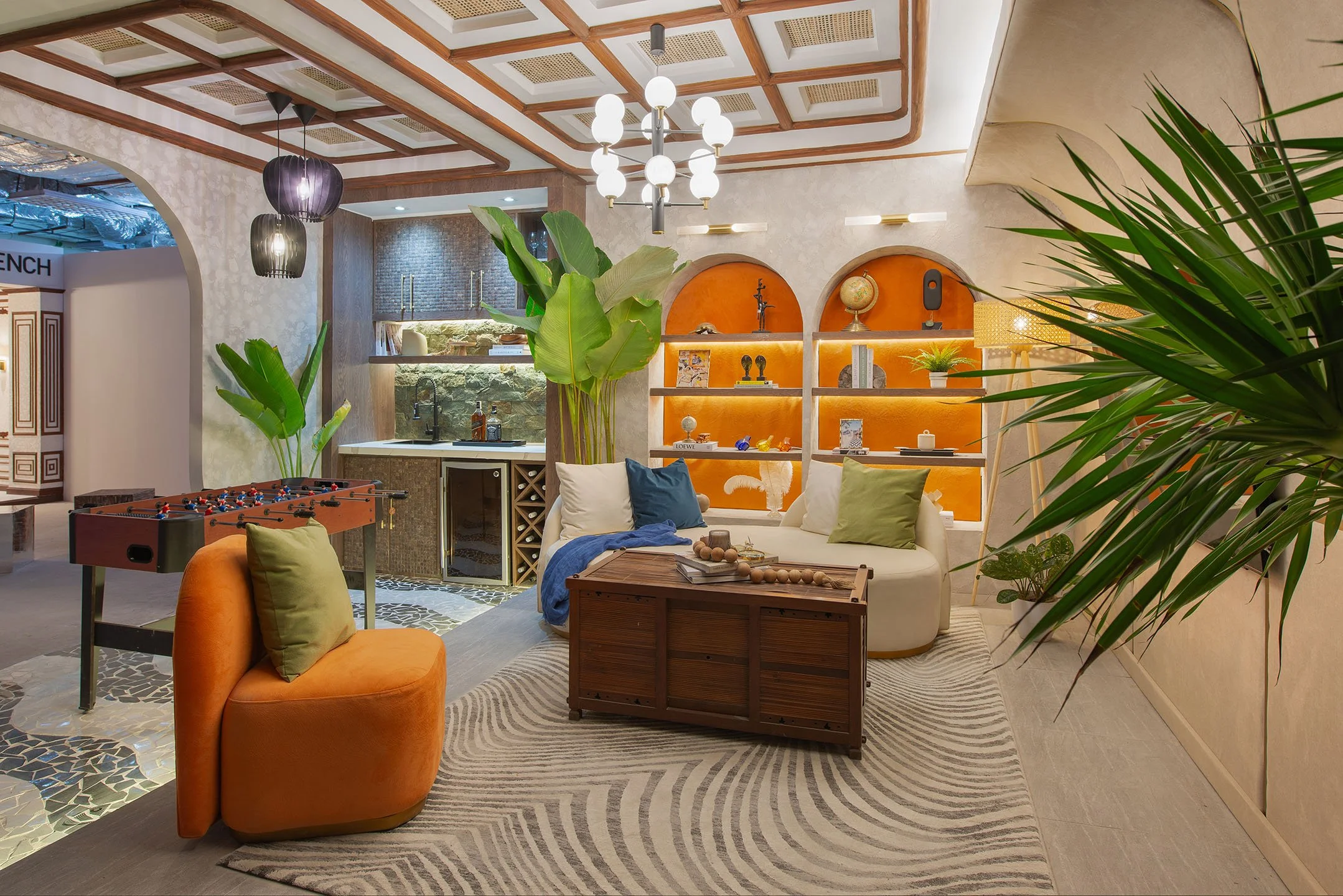 A cozy living room with a view into a small kitchen area, decorated with modern and tropical accents. It features a white curved sofa with colorful pillows, a wooden coffee table, large green plants, and a unique ceiling light fixture. The room has a patterned rug and a built-in shelf with decorative items and books.