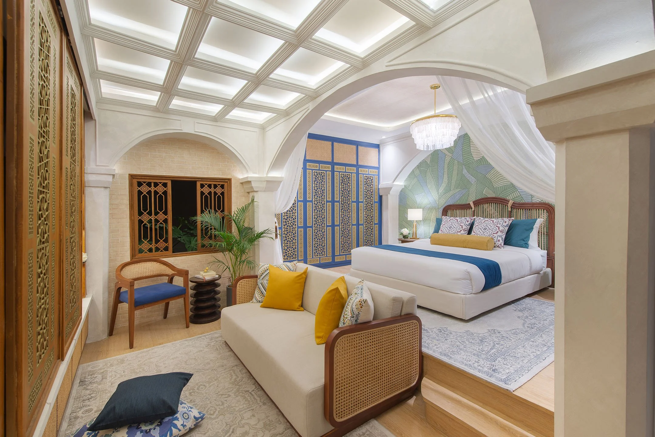 A bedroom with a coffered ceiling, a chandelier, a bed with decorative pillows, a blue and yellow color scheme, a sitting area with a vase and chairs, and tropical wallpaper.