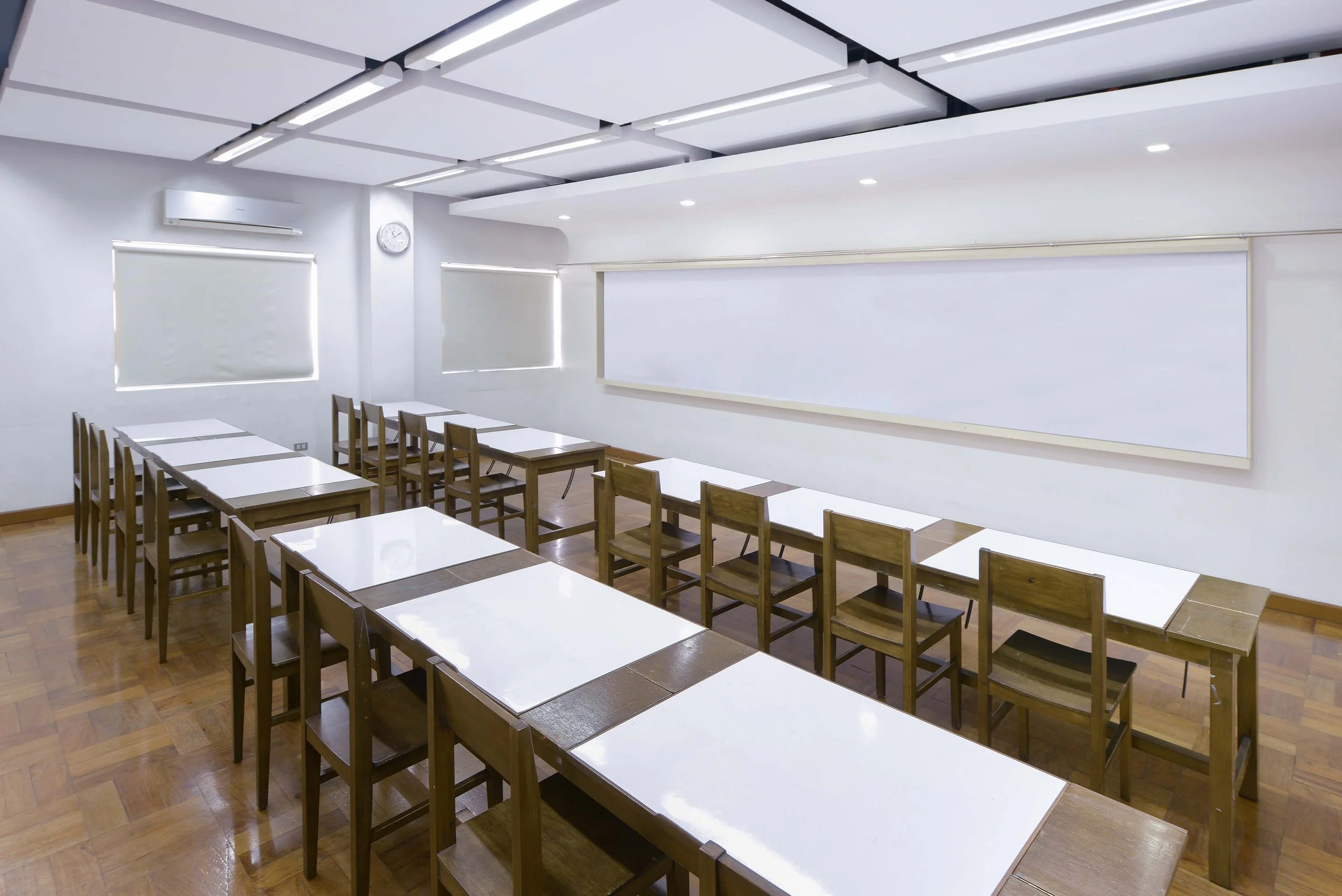 An empty classroom with white tables and wooden chairs, white walls, a whiteboard, a clock, and windows with blinds.