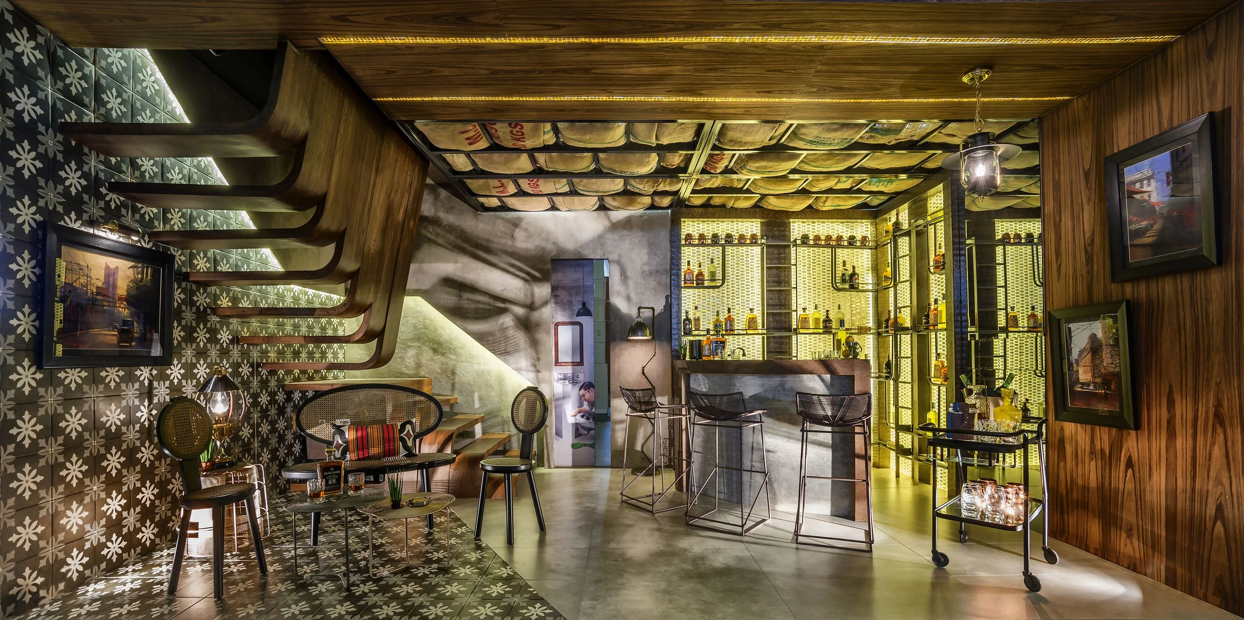A modern restaurant interior with patterned tile flooring, wooden accents, and a illuminated yellow shelving unit behind the bar. There are chairs, tables, and artworks on the walls, with a person sitting near the wall.