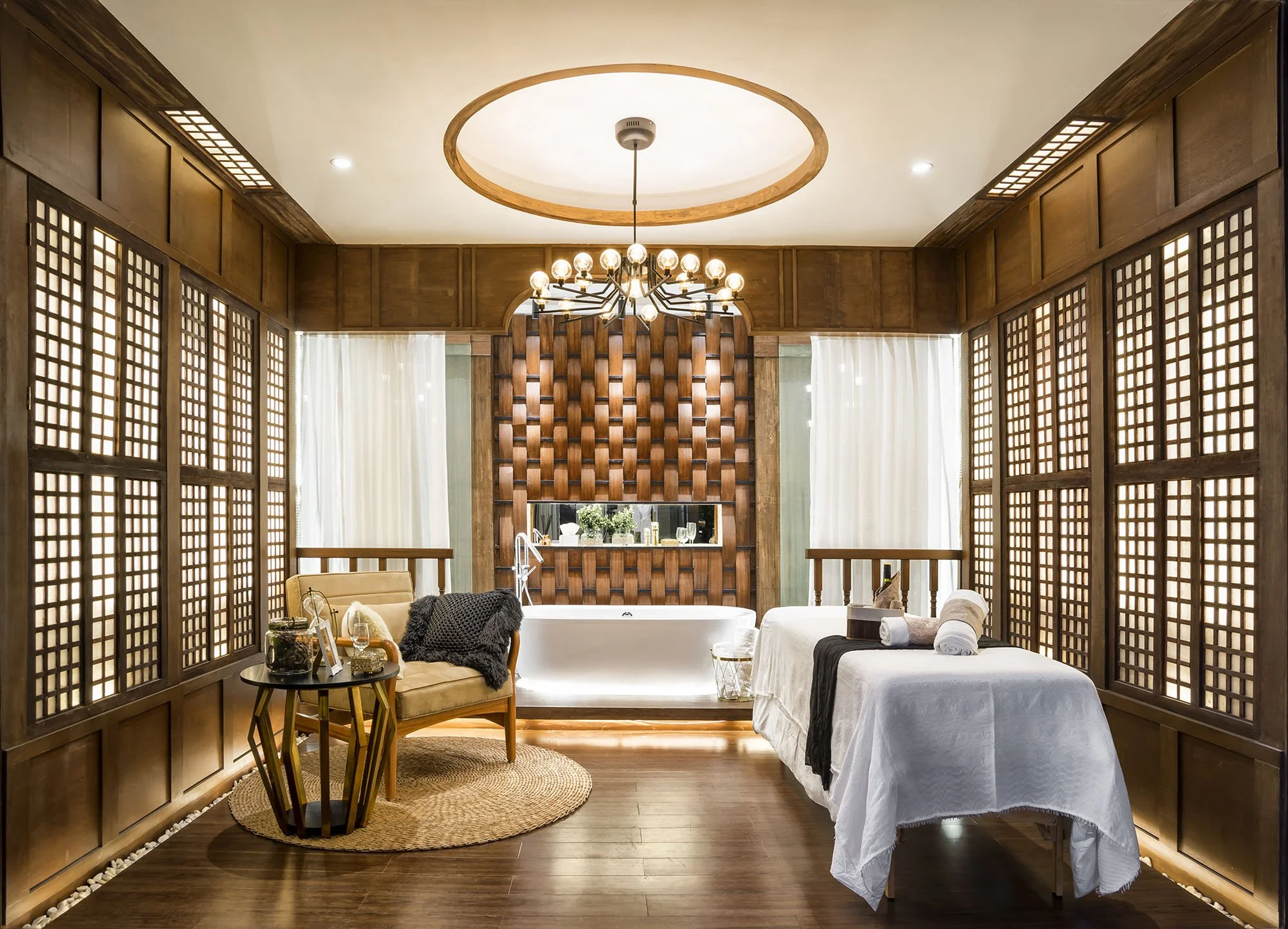 A room with wooden lattice dividers, a chandelier, a freestanding bathtub, and a bed with white linens, decorated in a modern rustic style.