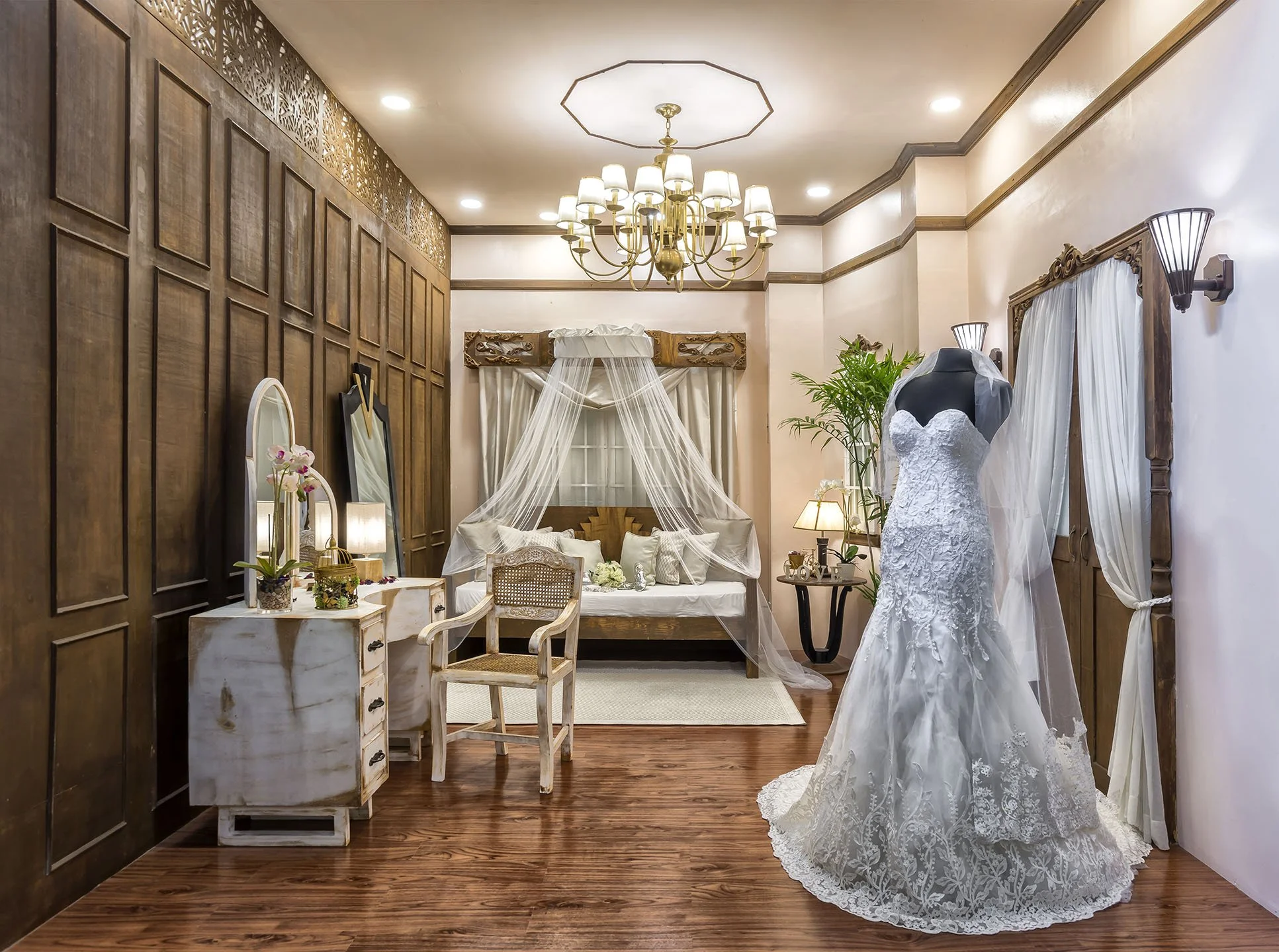 A bridal indoor showroom with a wedding dress on a mannequin, multiple tables and chairs, a chandelier, a wooden accent wall, and decorative plants.