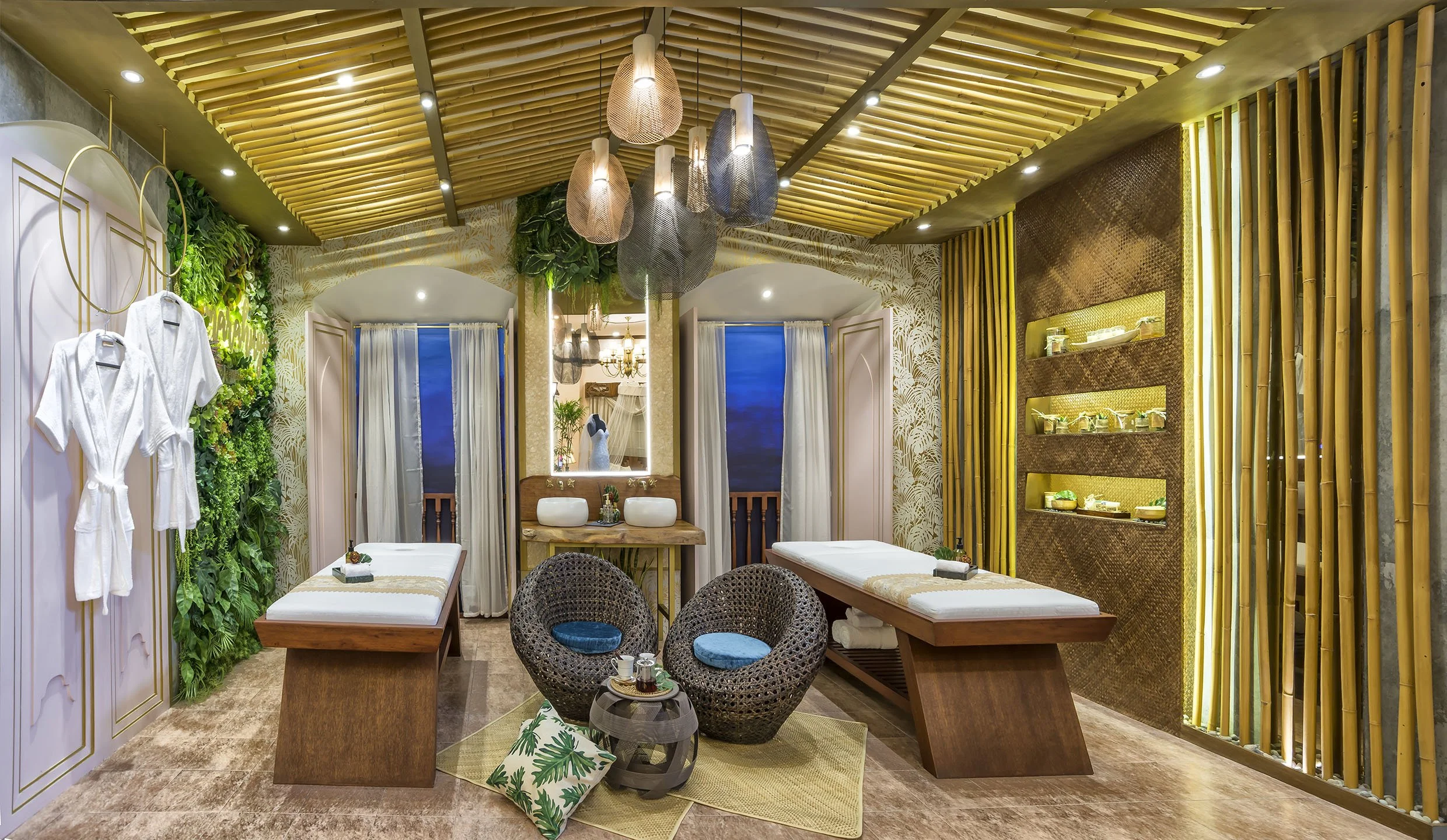 Spa treatment room with massage tables, wicker chairs, green wall with plants, hanging woven light fixtures, bamboo decor, and lush tropical accents.