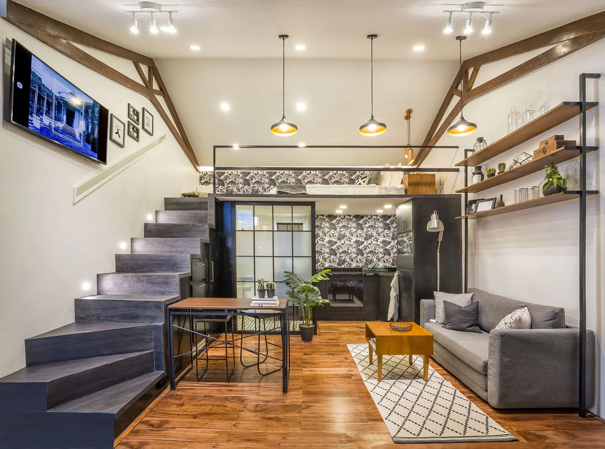 A modern, two-story living space with wooden floors, a gray sofa, and a loft bedroom above. The room features a flat-screen TV on the wall, bookshelves with decorative items, and a wood dining table with black wire chairs. There are hanging pendant lights and a staircase with dark wood steps.