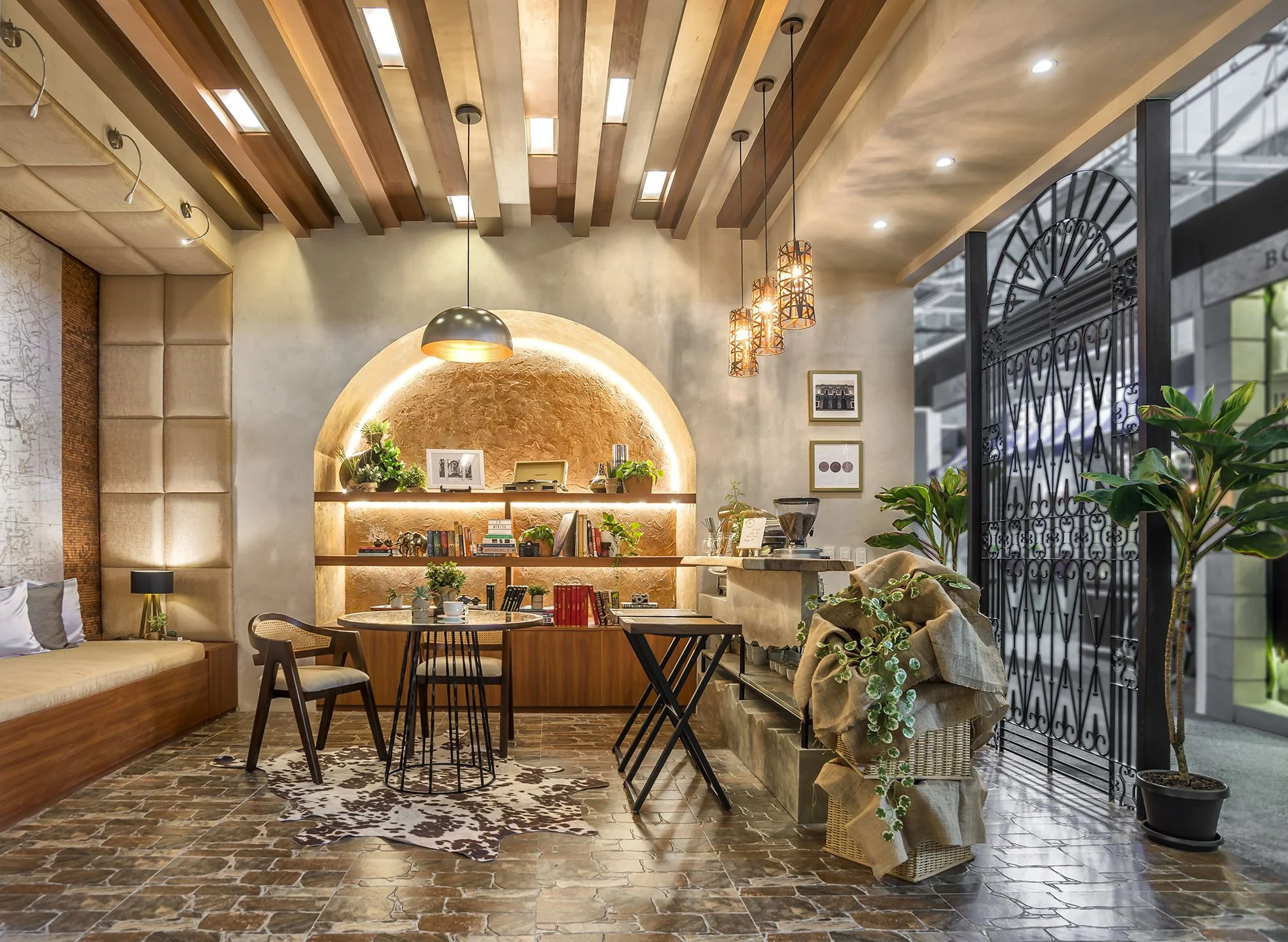 A cozy indoor seating area with a built-in cushioned bench along the left wall and a playful patterned rug on a stone-tile floor. There is a wooden wall with hanging lights and a round table with four chairs in the center. The back wall features a decorative arc with plants, framed pictures, and shelves, and the ceiling has wooden beams with hanging pendant lights. To the right, a large window with a black wrought-iron gate and lush green plants adds natural light.