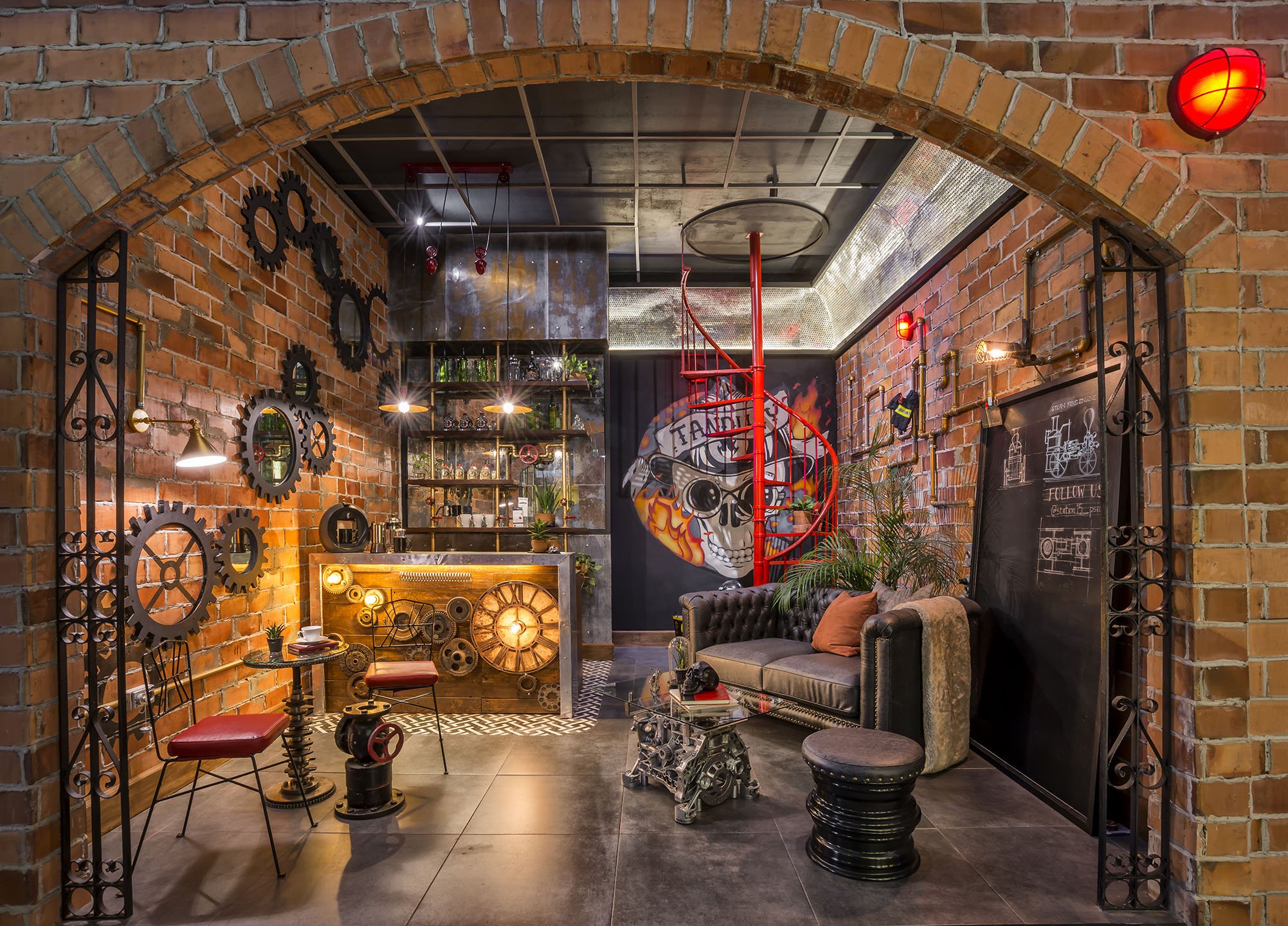 Interior of a room with brick walls, industrial decor, and vintage furniture including a black leather sofa with orange pillows, a glass coffee table with mechanical parts, and a small accent table with a plant and tableware. The walls are decorated with gear-shaped mirrors, and there is a red spiral staircase leading up. The ceiling has a mix of exposed concrete and metal grid pattern, and there are wall-mounted red lights and a graffiti-style mural of a skull with flames.