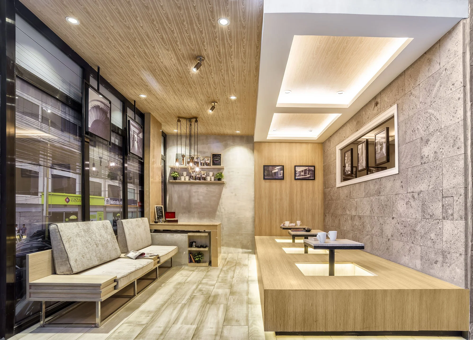 Interior of a modern cafe with wooden and stone walls, ceiling lights, a window with blinds, and seating with beige cushions. There are framed pictures on the walls and a small shelf with decor items and potted plants.