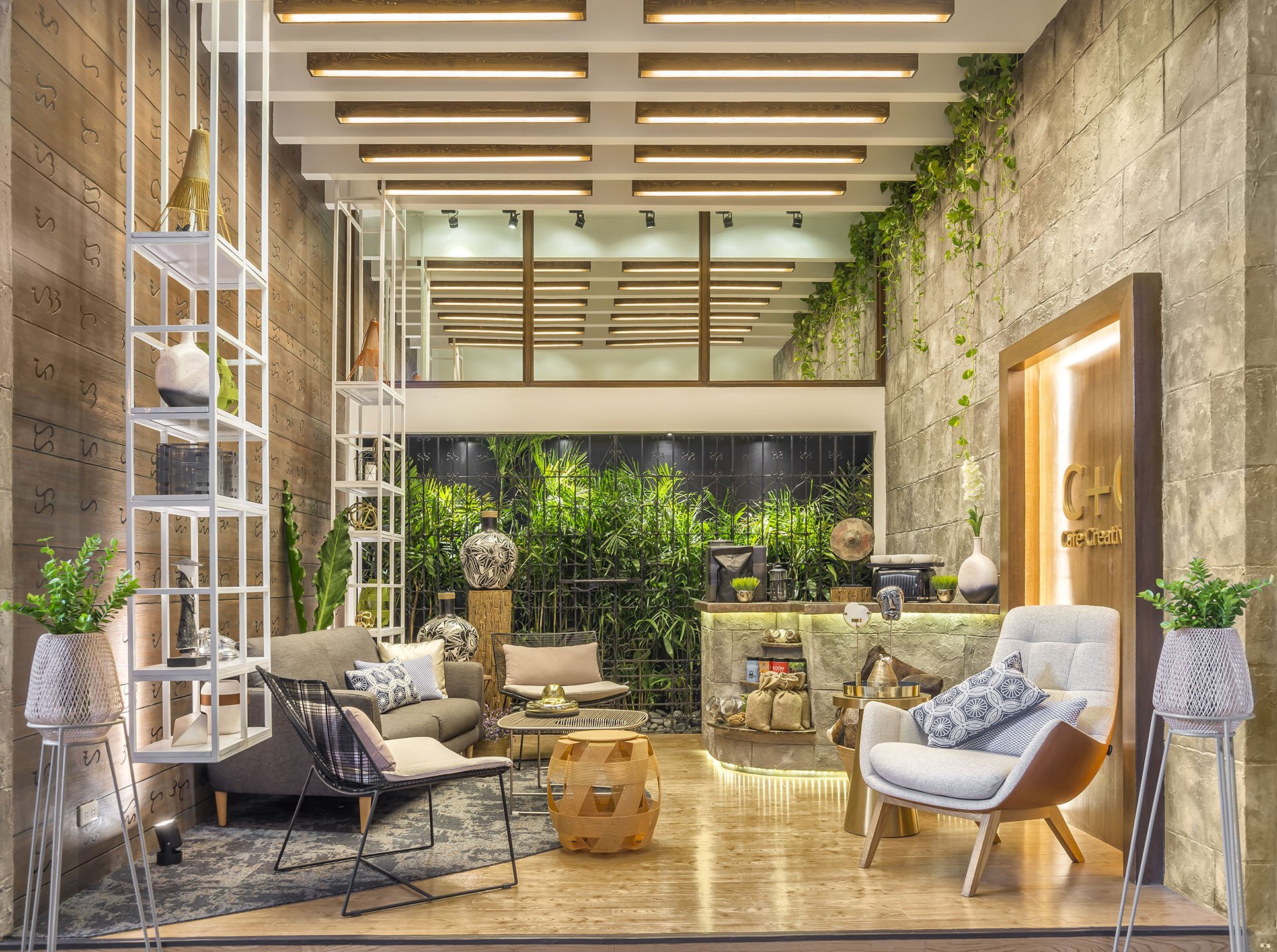 Modern lobby area with various couches, armchairs, and decorative pillows. There are plants along a back wall and shelving on the left with vases and sculptures. The ceiling has wooden beams with lighting, and the floor is wood with a patterned rug. Signage on the right shows 'C + C Cafe Creative'.