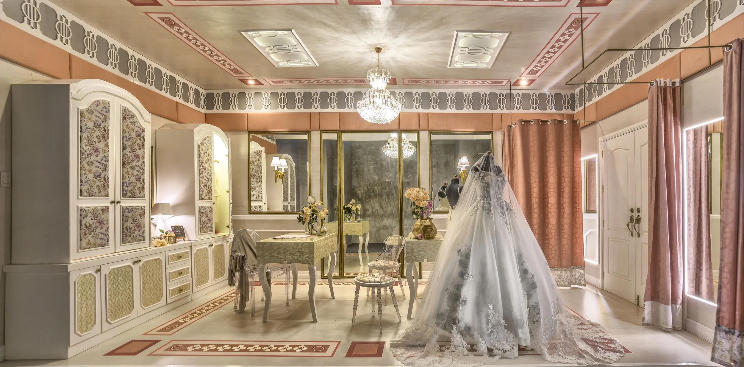 A bridal salon with a wedding dress on a mannequin, chandeliers hanging from the ceiling, pink curtains, floral wallpaper, a mirror, and vintage-style furniture.