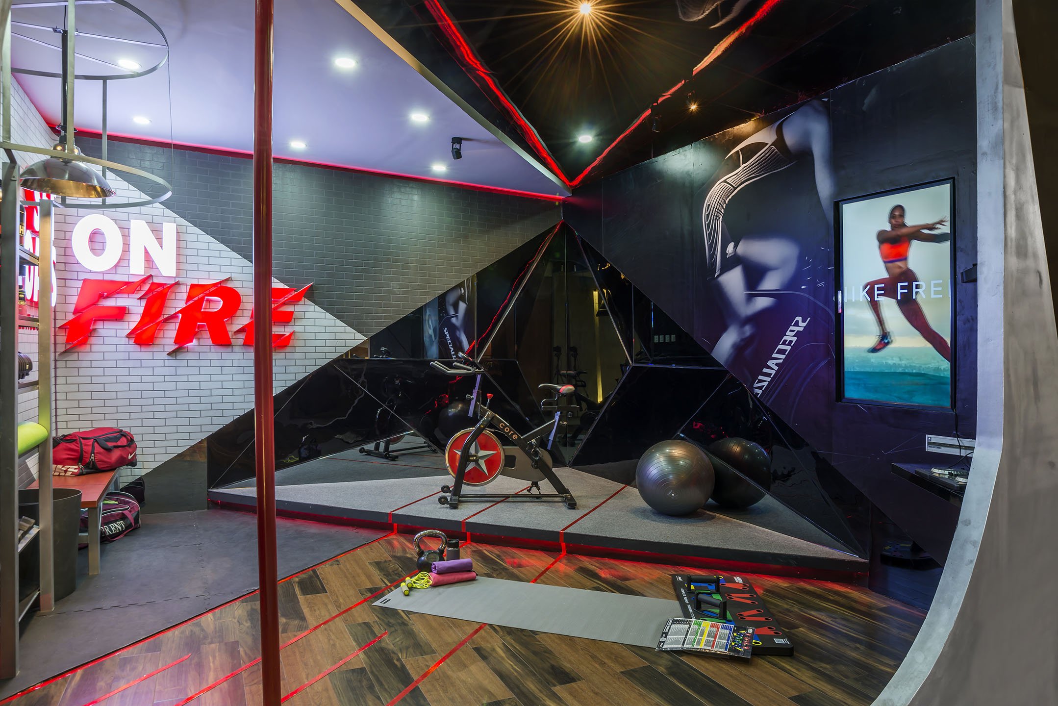 A modern gym with workout equipment, a stationary bike, exercise balls, and towels on the floor. The walls feature bright neon signs and large digital screens displaying activewear advertisements. There are glossy black and dark gray walls with red accent lighting.