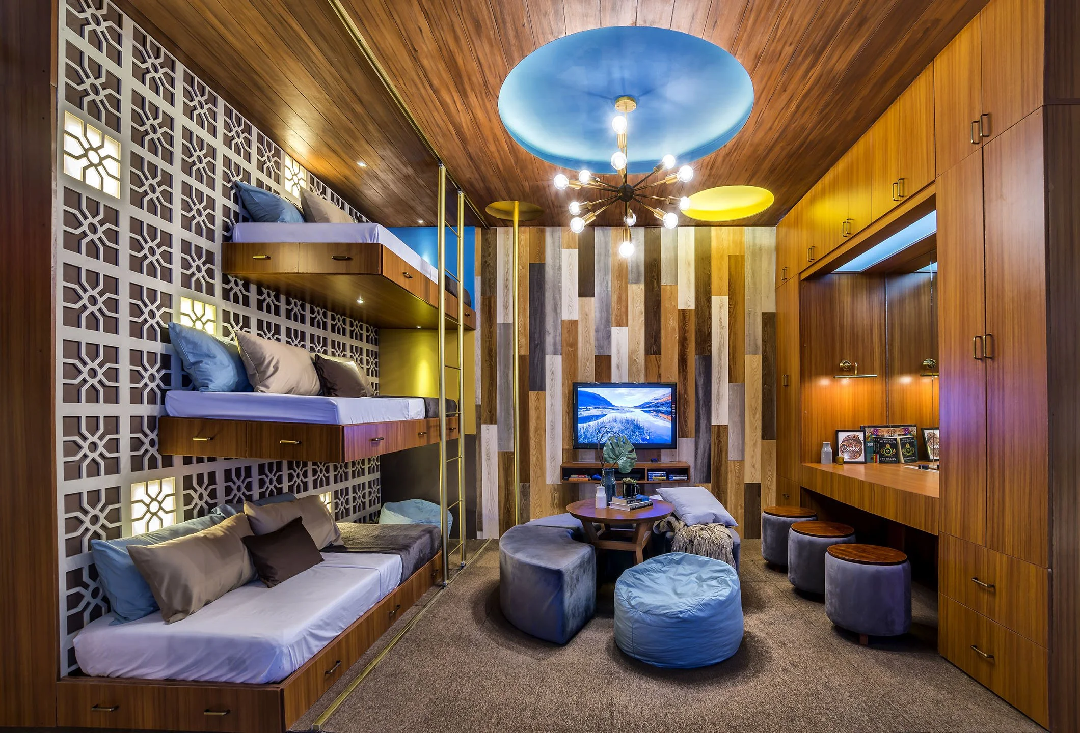 A cozy living space with built-in wooden beds, colorful light fixtures on the ceiling, and a small television. The room features a wood-paneled ceiling, a decorative wall with lighting, and a mix of furniture including bean bags and a round table.