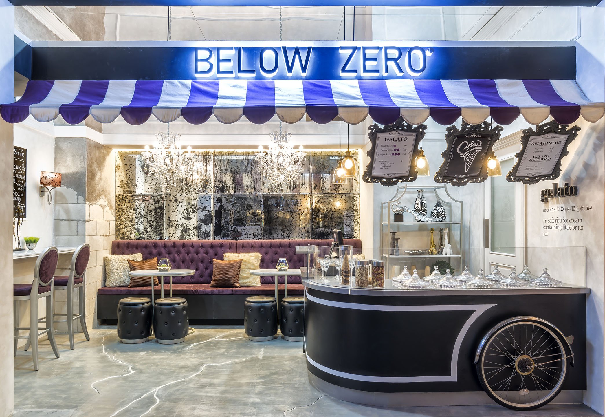 Ice cream shopfront with a purple and white striped awning and blue illuminated sign that says "Below Zero." Inside is a cozy seating area with purple tufted banquette, pillows, and ornate chandeliers, with a menu board displaying gelato options and a black and white ice cream cart in the foreground.