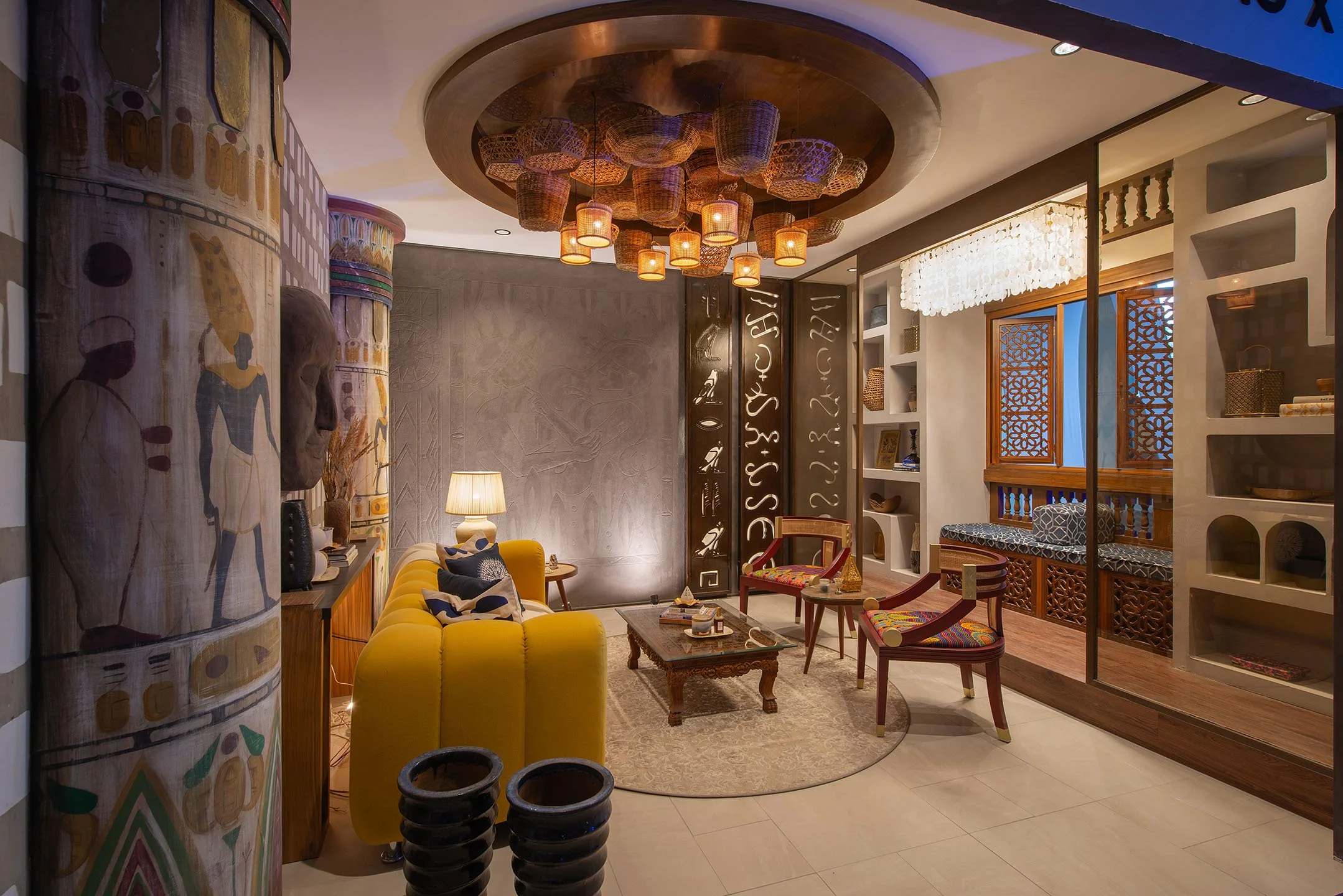 Interior of a living room with a yellow sofa, wooden coffee table, patterned chairs, shelves, and a decorative ceiling with hanging baskets and warm lighting.