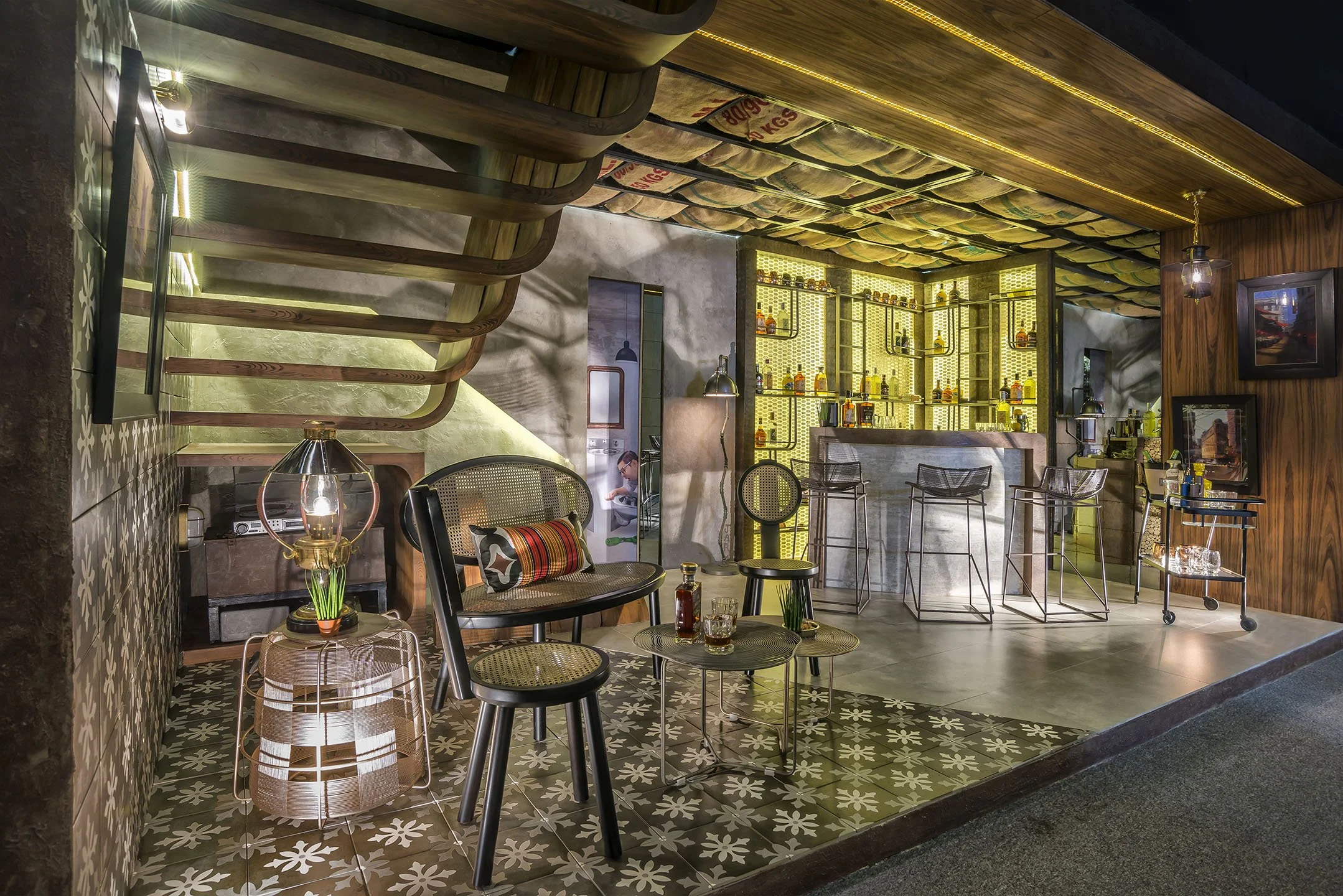 Interior of a modern restaurant or cafe with wooden accents, patterned tile floors, and a liquor shelf display with yellow backlighting. There are chairs, small tables, and decor including lamps and framed pictures.