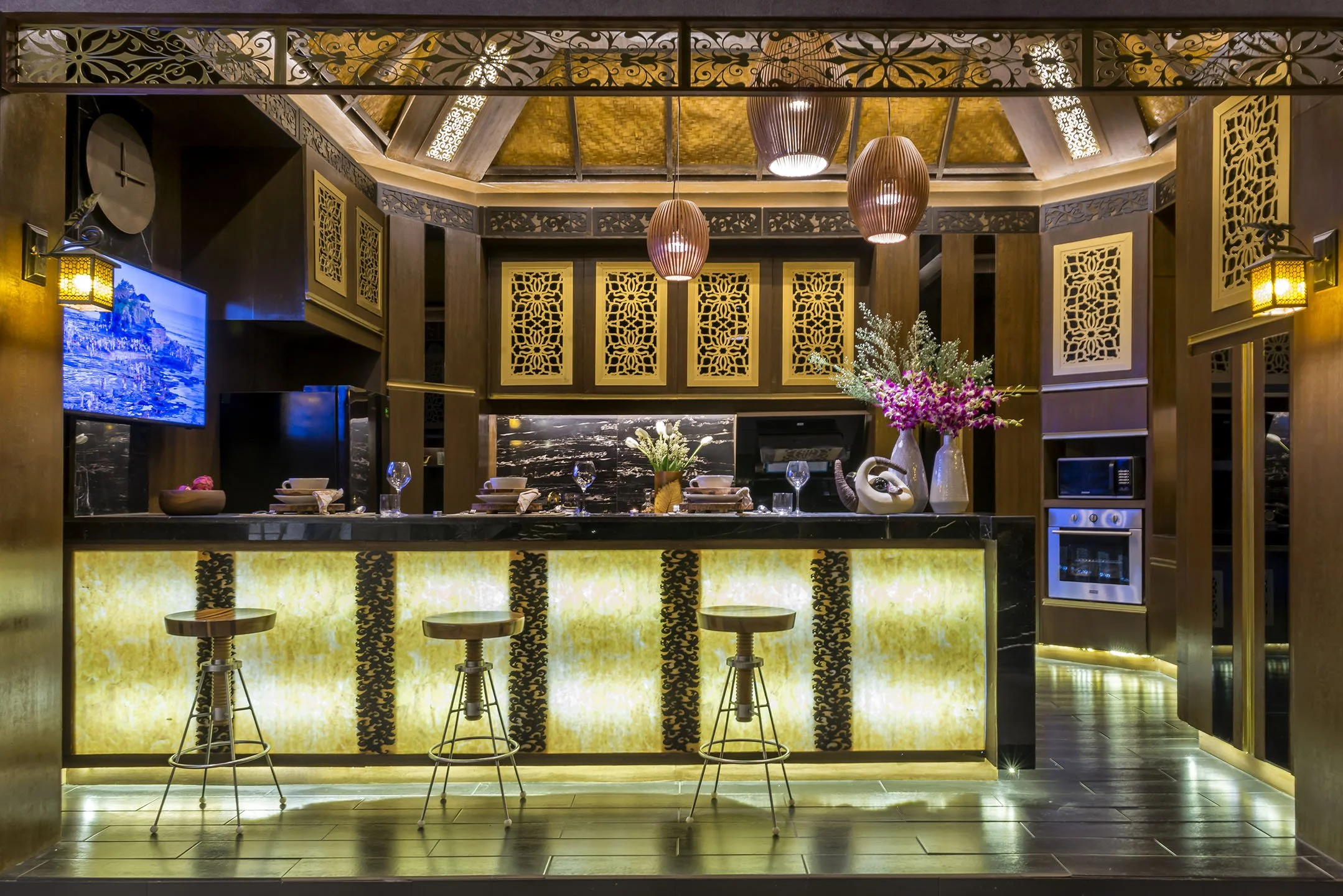 Elegant restaurant interior with a bar counter, floral arrangements, hanging pendant lights, and decorative metalwork panels on the walls.