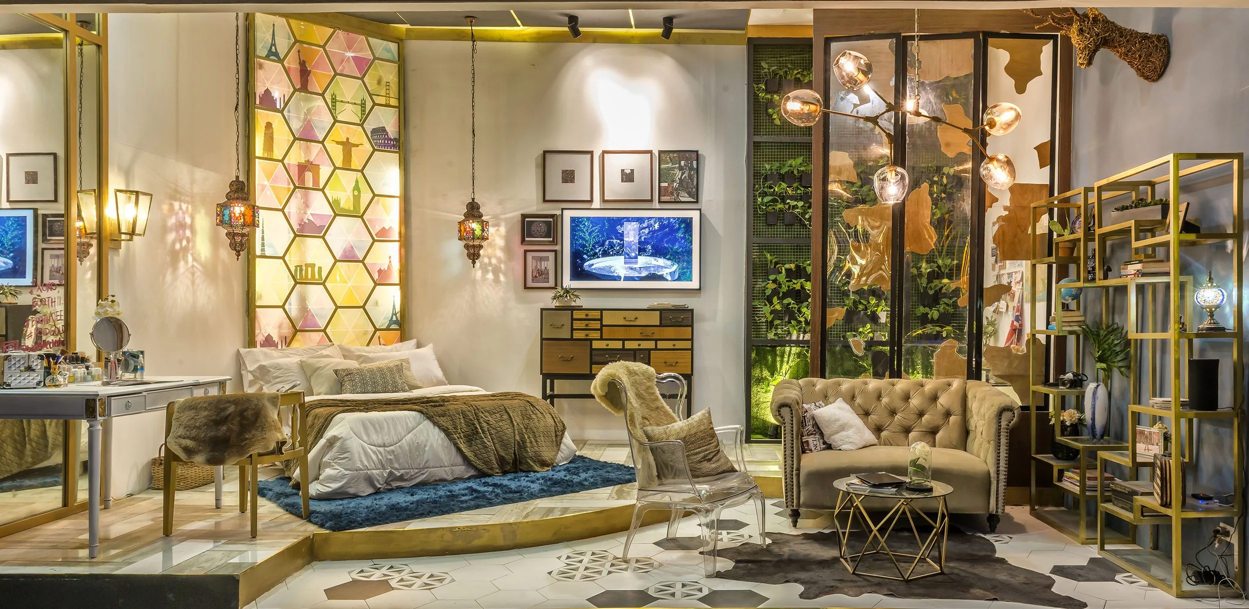 A cozy living space with a bed, a sofa, and various decorative elements including hanging lanterns, framed artwork, a television, and a wall of greenery.