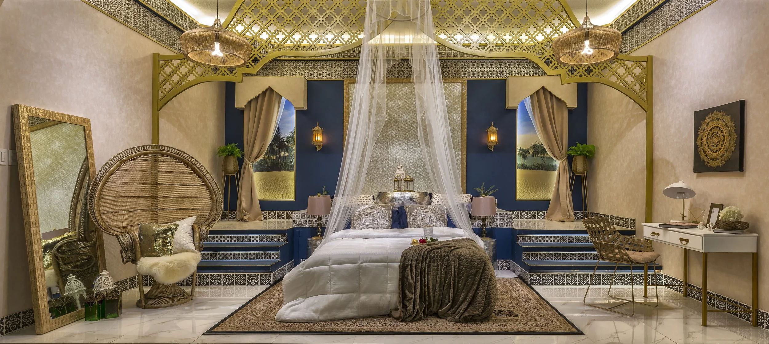 A luxurious bedroom with beige walls and intricate gold and blue accents. The bed has white bedding, decorative pillows, and a canopy of sheer fabric. There are matching blue nightstands with lamps, two gold lanterns, a decorative mirror, a chair with a pillow, and a small white desk with a chair and accessories. Two large paintings of a desert landscape are behind the bed, and the ceiling has ornate gold detailing with hanging lights.