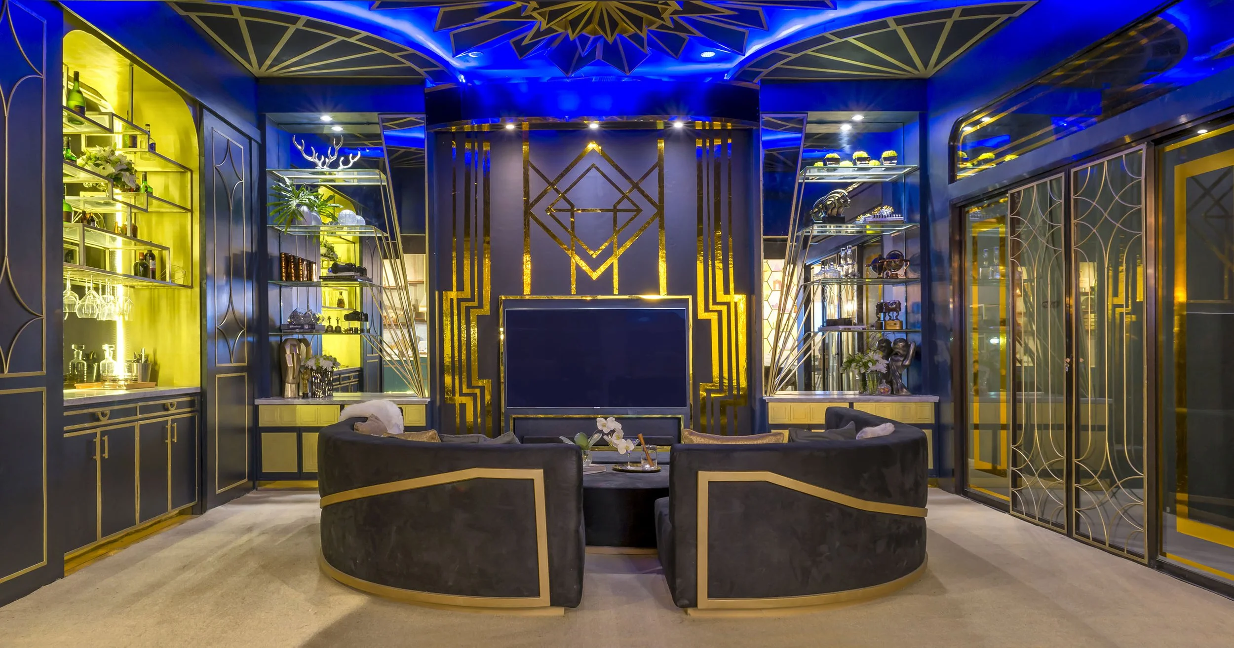 Luxurious living room with blue and gold decor, black couches, built-in cabinets with lighting, decorative plants, and a large flat-screen TV surrounded by a gold geometric frame.