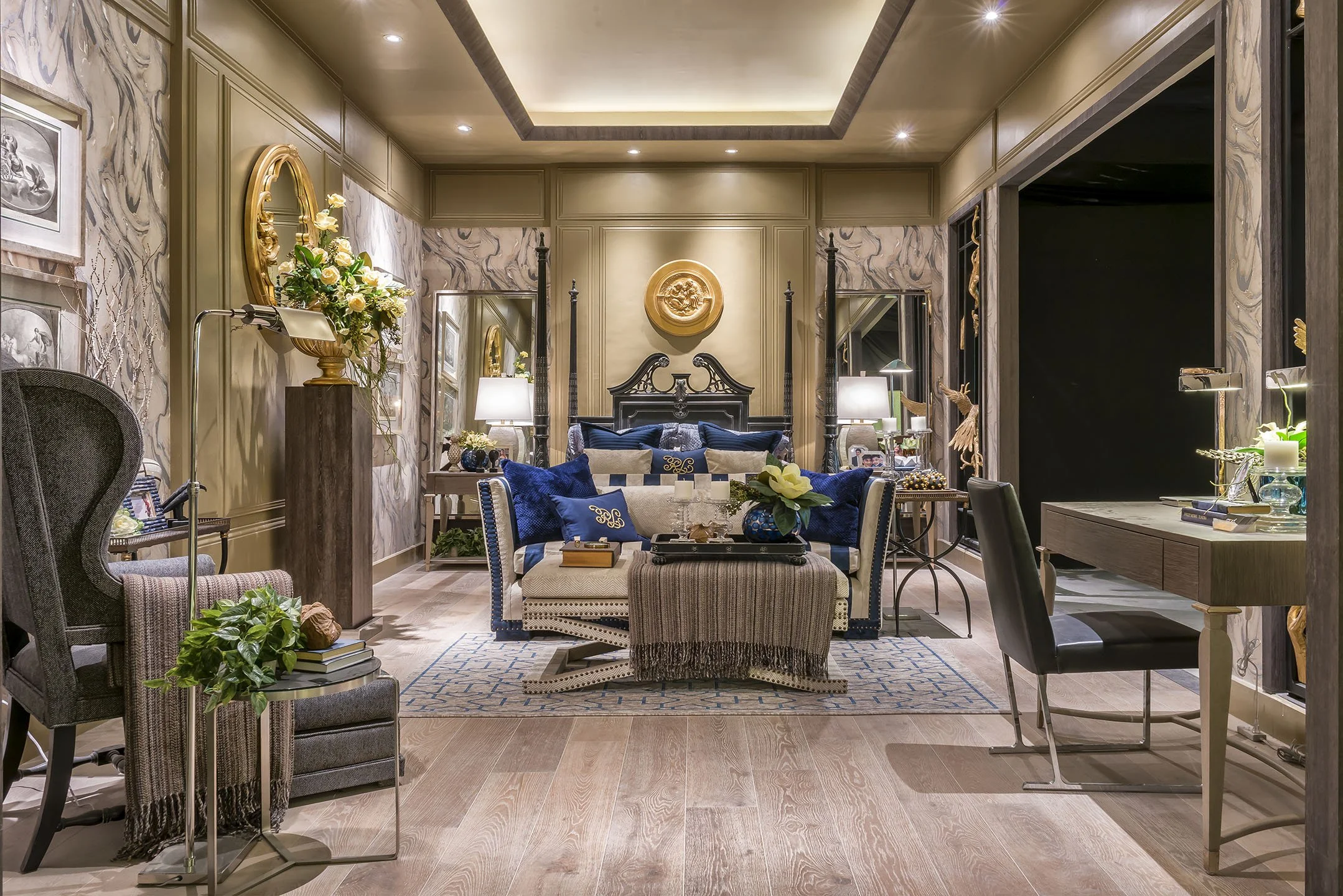 Interior of a living room with beige walls, marble accents, a four-poster bed with a dark headboard, blue and beige cushions, a patterned area rug, small tables with lamps, flowers, and decorative objects, and framed art on the walls.
