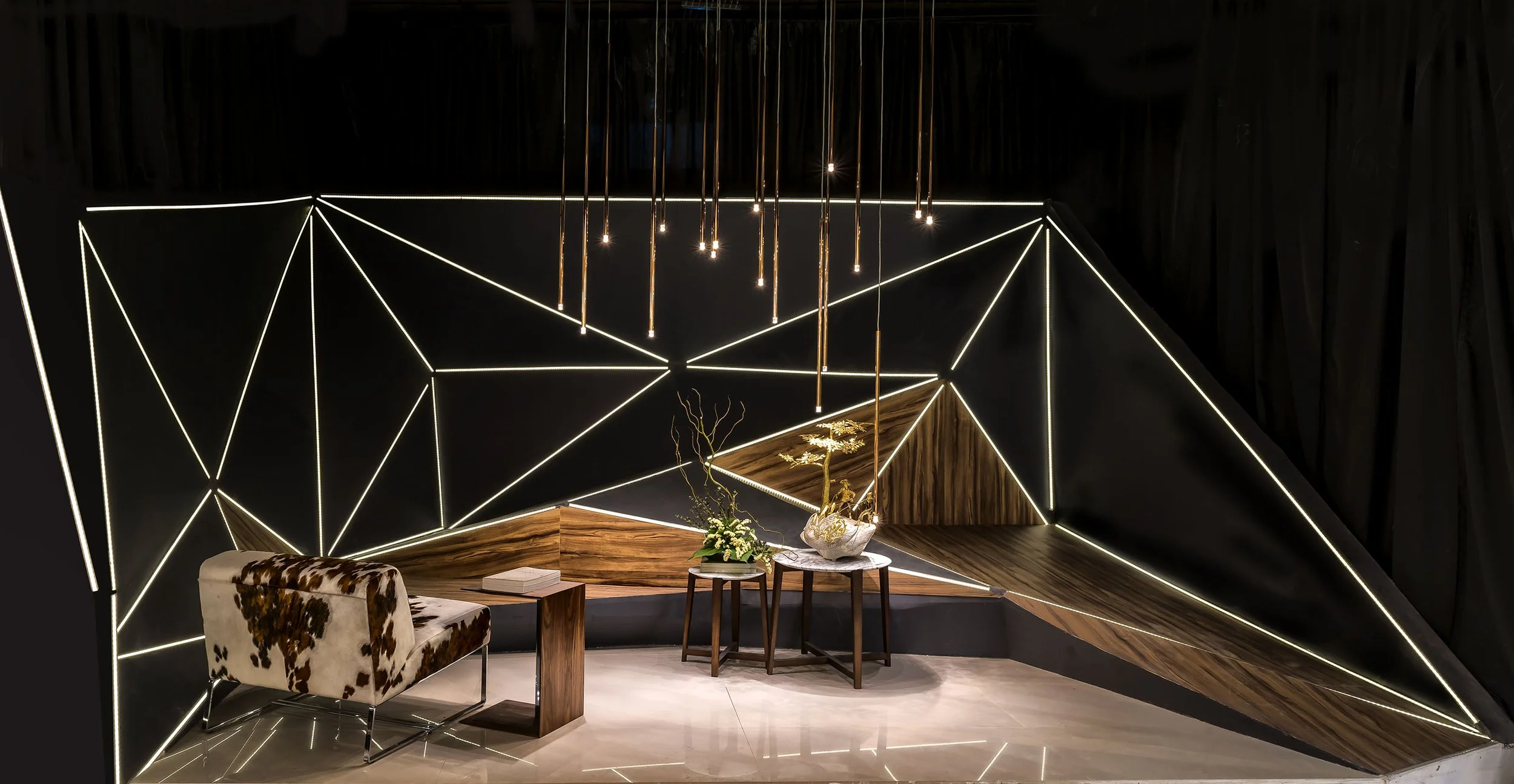 Modern interior with geometric black wall adorned by white LED strip lights, a cowhide upholstered chair, a wooden table with a vase of flowers, and a black curtain in the background.