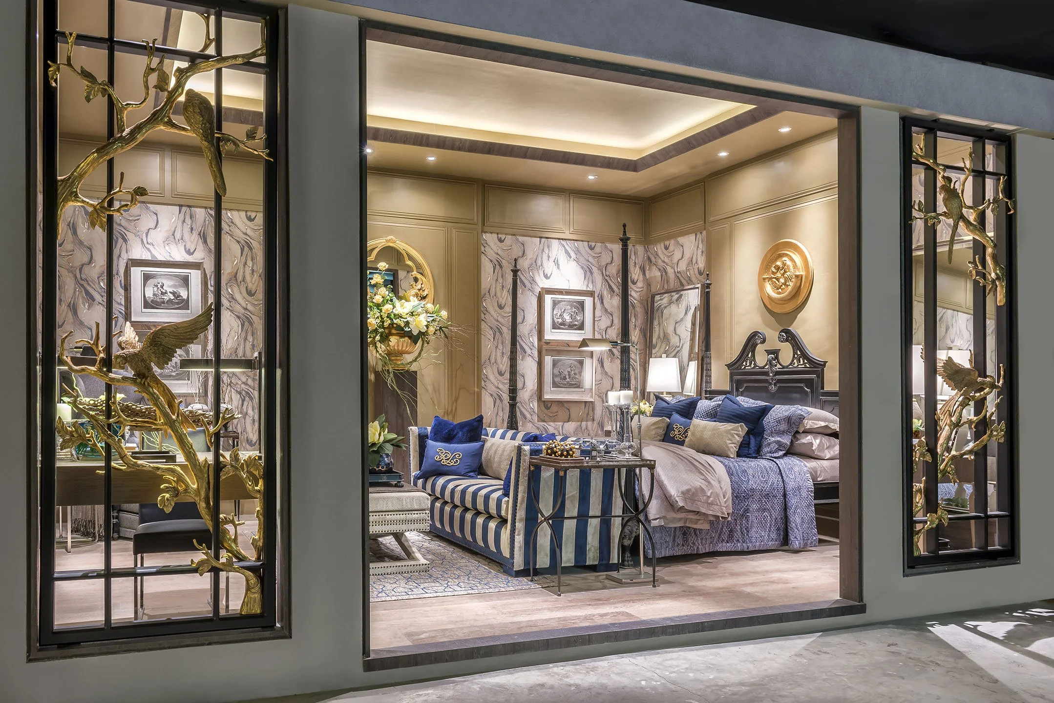 A luxurious bedroom with cream-colored paneled walls, framed artwork, and elegant gold wall decor. The room features a dark wooden four-poster bed with a carved headboard, draped with blue and beige linens and decorative pillows. A blue and white striped sofa with matching pillows is positioned near a side table with floral arrangements and lamps. The ceiling has recessed lighting and cove lighting that highlights the layered ceiling design.