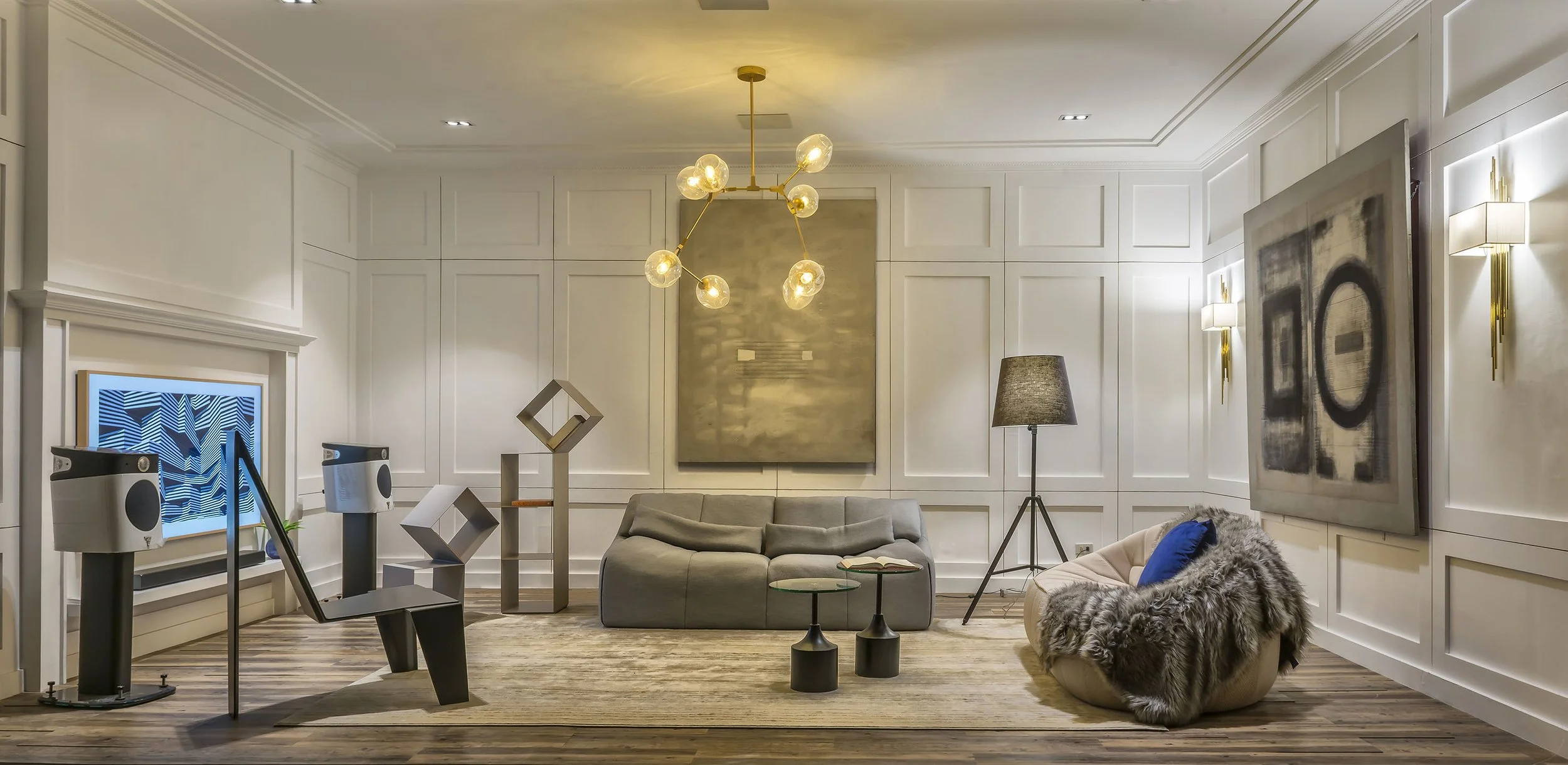 Modern living room with white paneled walls, gold chandelier, gray sofa, abstract art, a floor lamp, and mounted flat-screen TV with geometric patterns.