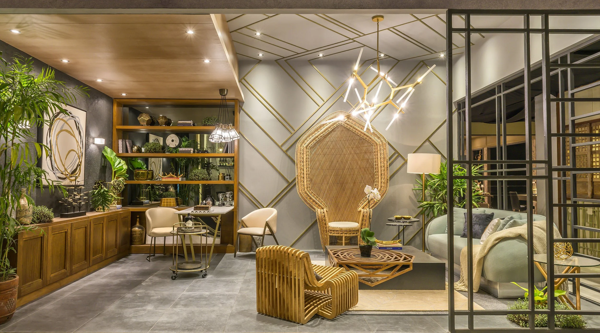 Modern interior living room with wooden shelves, lush green plants, a decorative wall with gold lines, a large wicker-backed chair, and a sofa, illuminated by warm lighting and artistic fixtures.