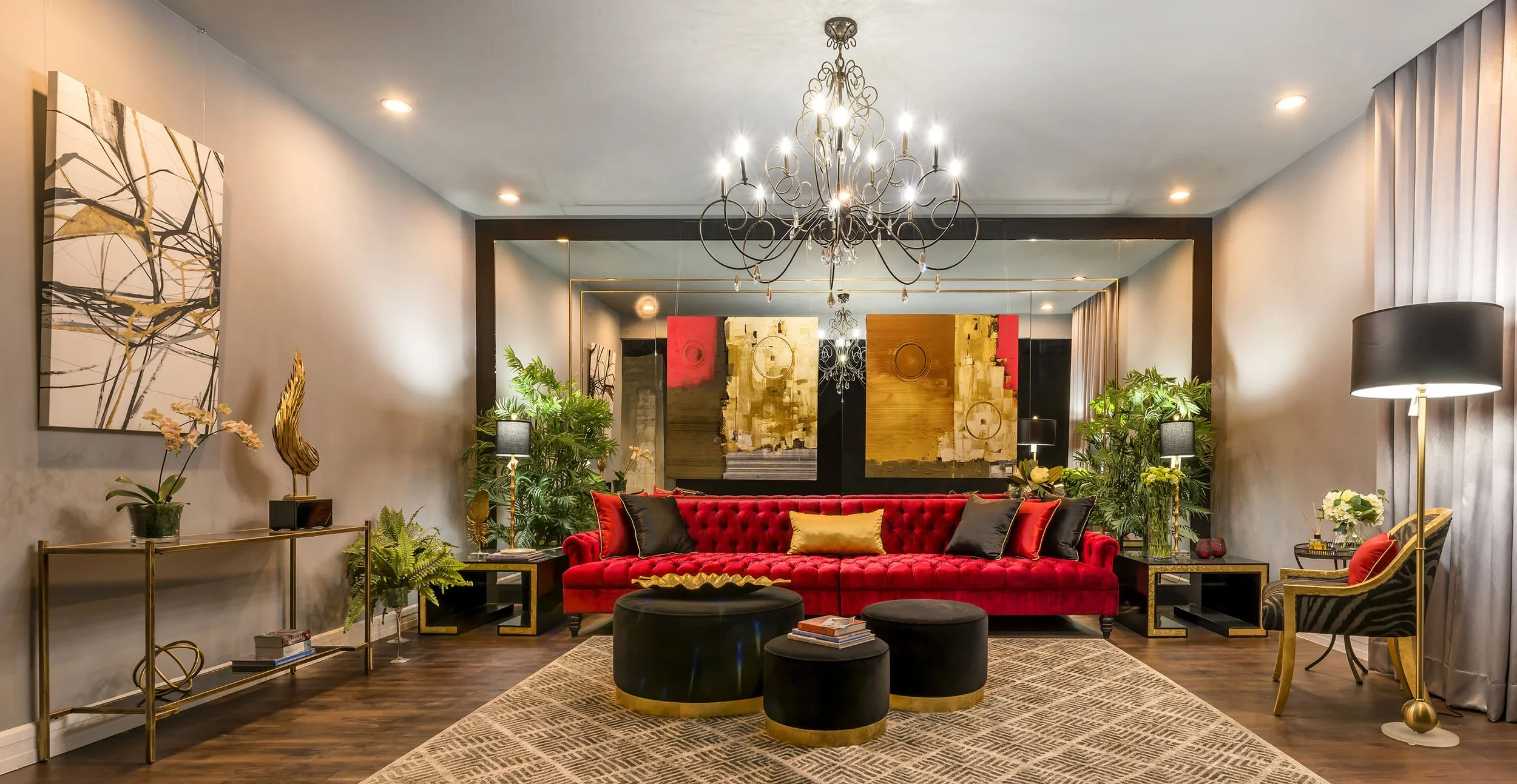 Living room with a bright red velvet sofa, black and gold ottomans, and various plants. Features a chandelier, abstract wall art, and gold accents.
