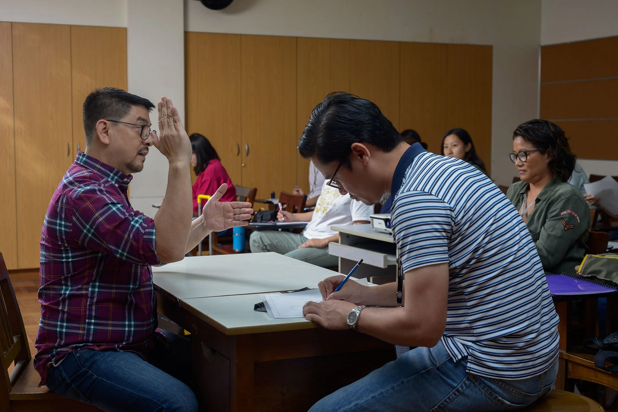 A man in a plaid shirt and glasses appears to be explaining something to another man in a striped shirt and glasses, who is taking notes. They are seated at a table in a classroom or meeting room with other people in the background.