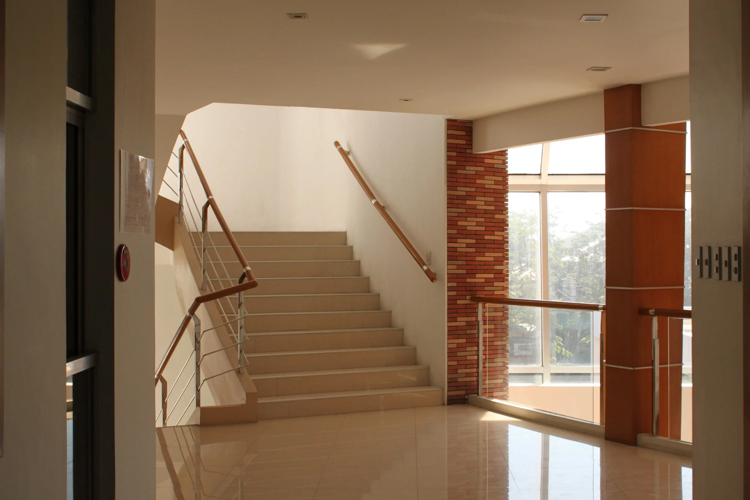 Interior of a modern building with stairs leading upward, large windows allowing natural light, and brick accent walls.
