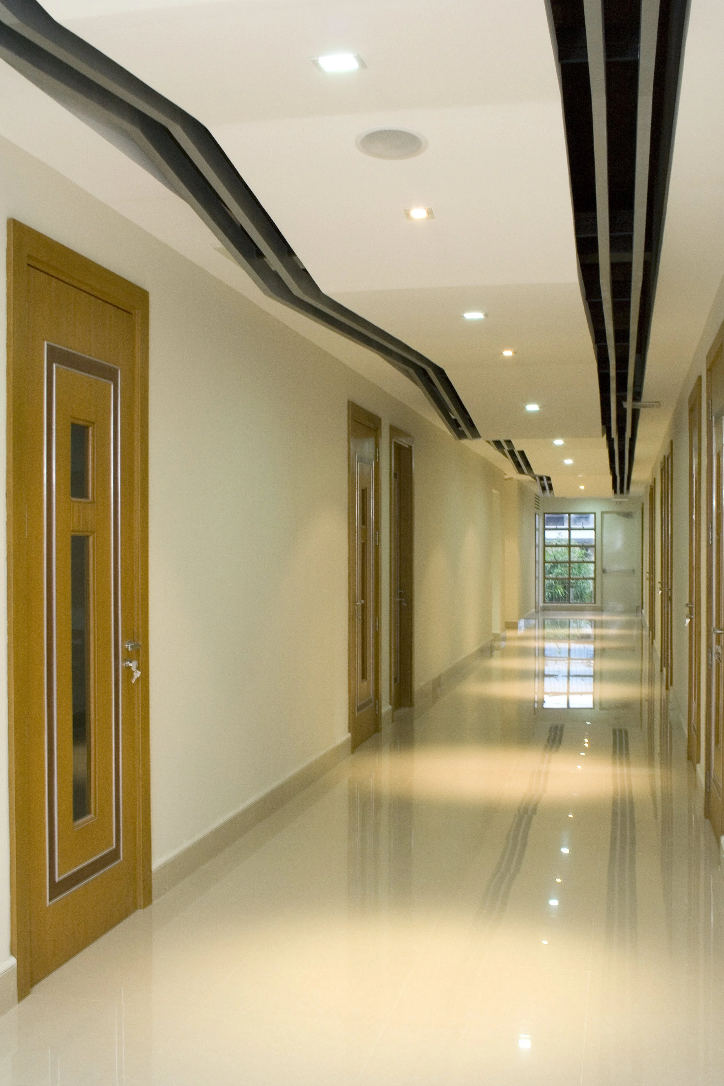 A long corridor with polished tile floor, cream-colored walls, wood framed doors on the left, and large windows at the end letting in natural light.