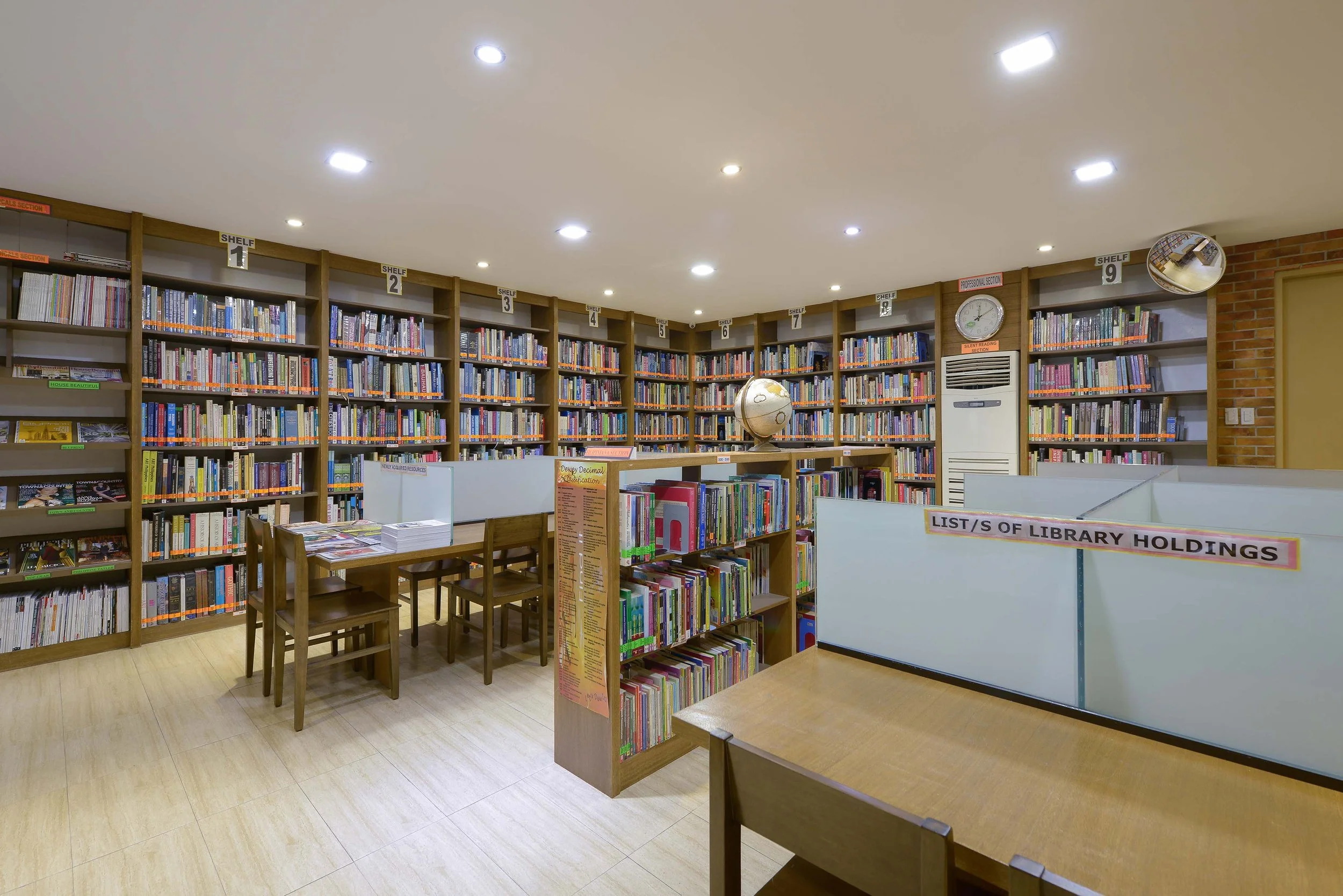 Interior view of a library with multiple bookshelves filled with books, a globe, a clock, and signs indicating different sections.