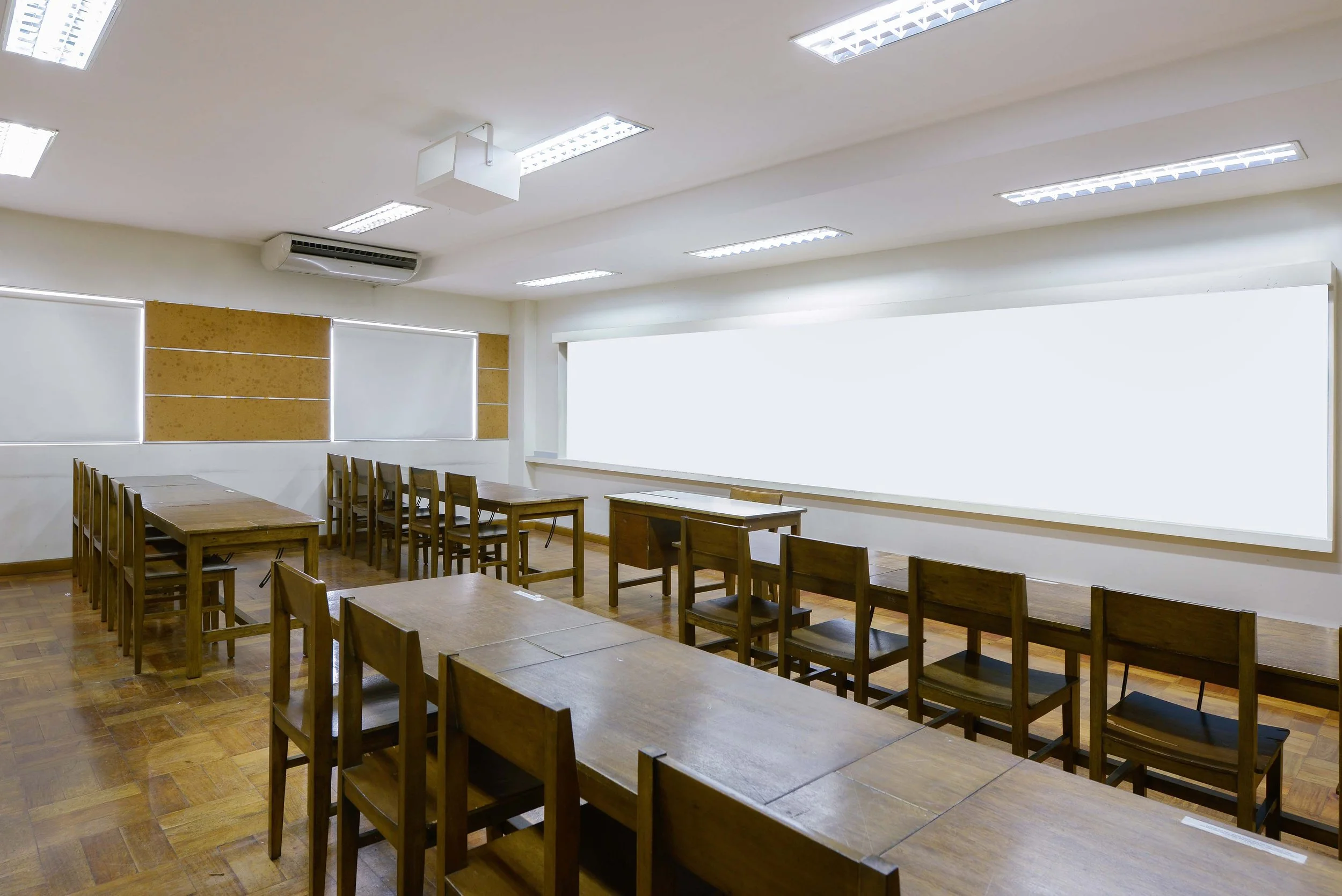 Classroom with wooden tables and chairs, whiteboard, window blinds, and ceiling lights.
