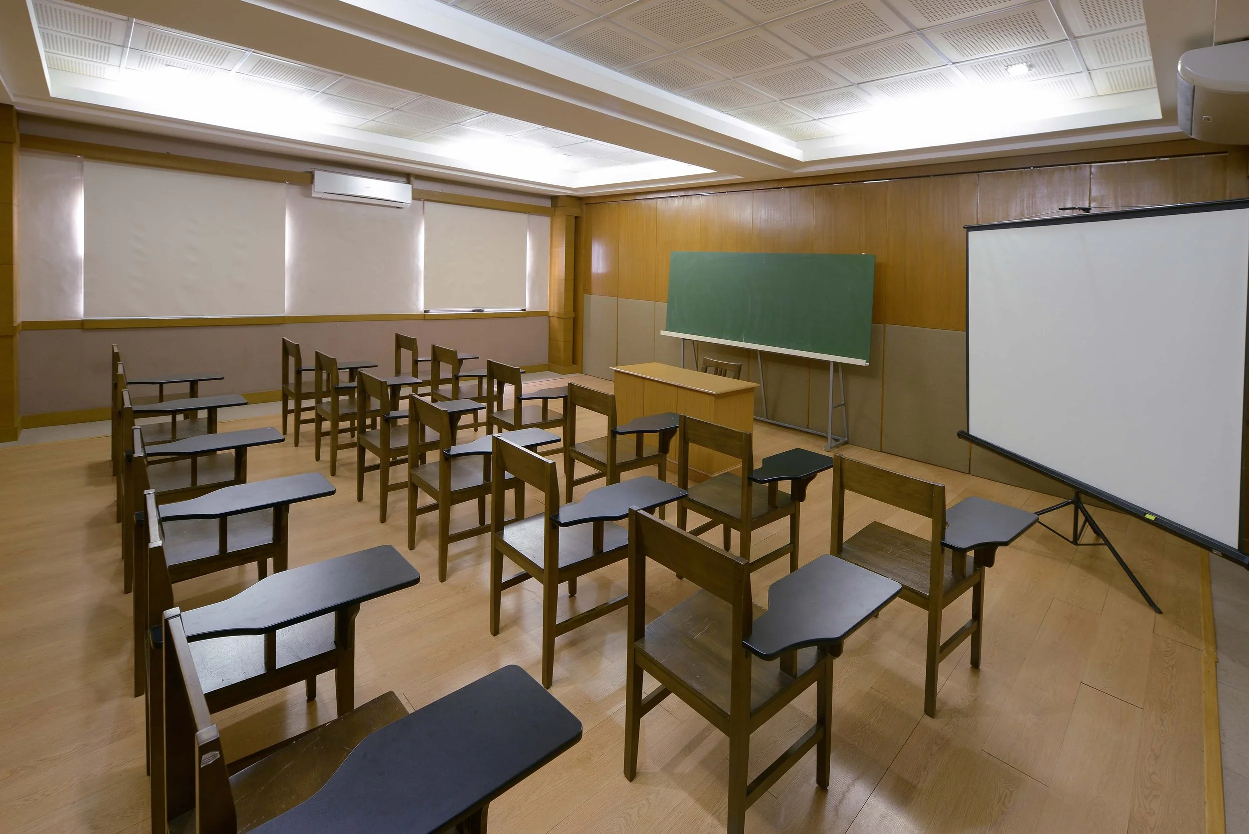 Empty classroom with wooden desks and chairs, a green chalkboard, a projector screen, wooden panel walls, and windows with blinds.