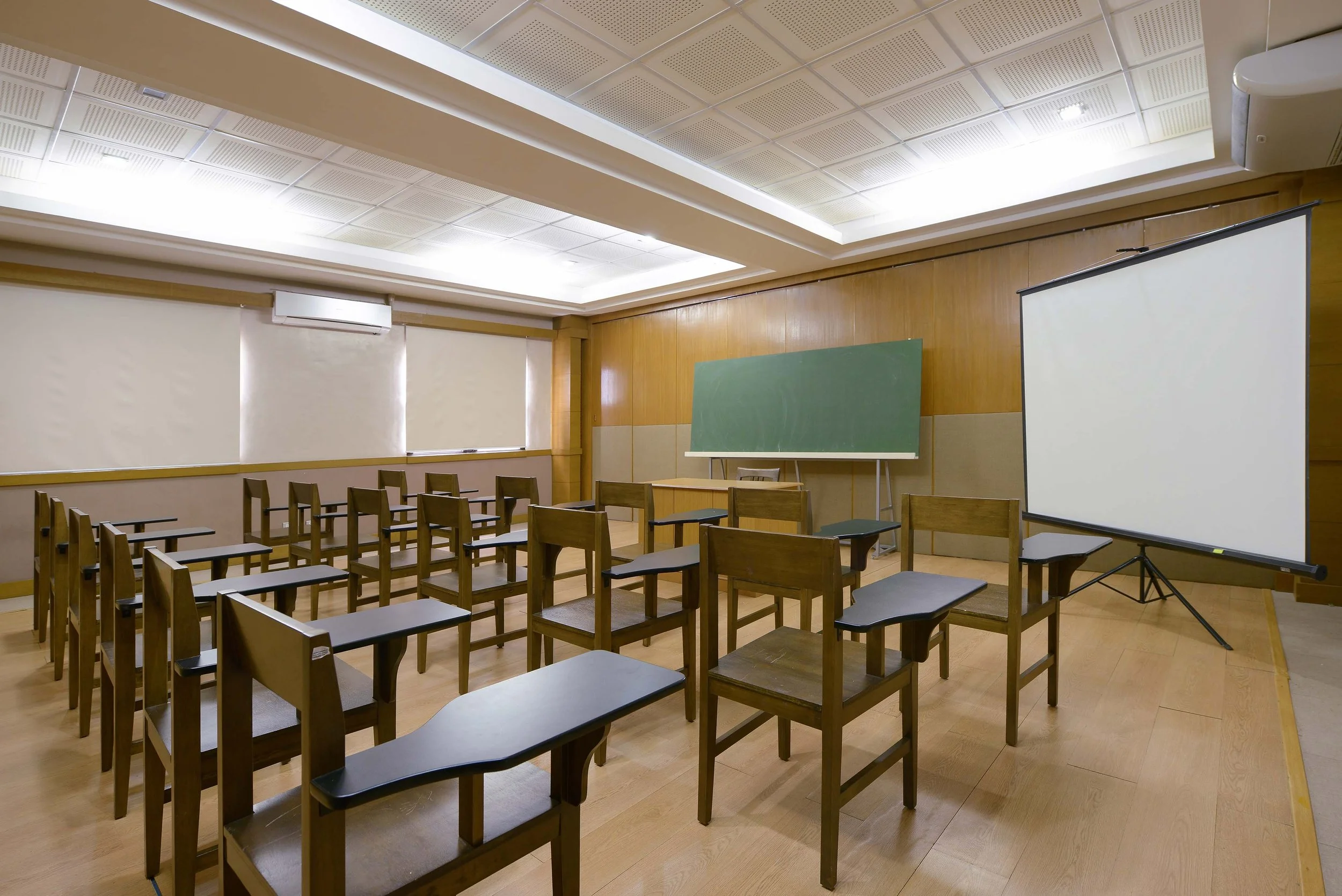 Empty classroom with rows of wooden chairs and tables, a green chalkboard, a projection screen, and windows with blinds, in a well-lit room with a drop ceiling.