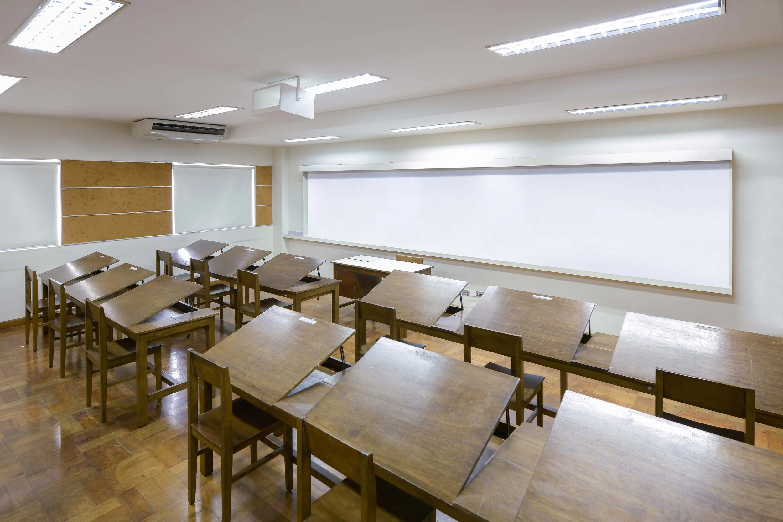 Empty classroom with wooden desks and chairs, white walls, a large whiteboard, and window blinds. Fluorescent ceiling lights.
