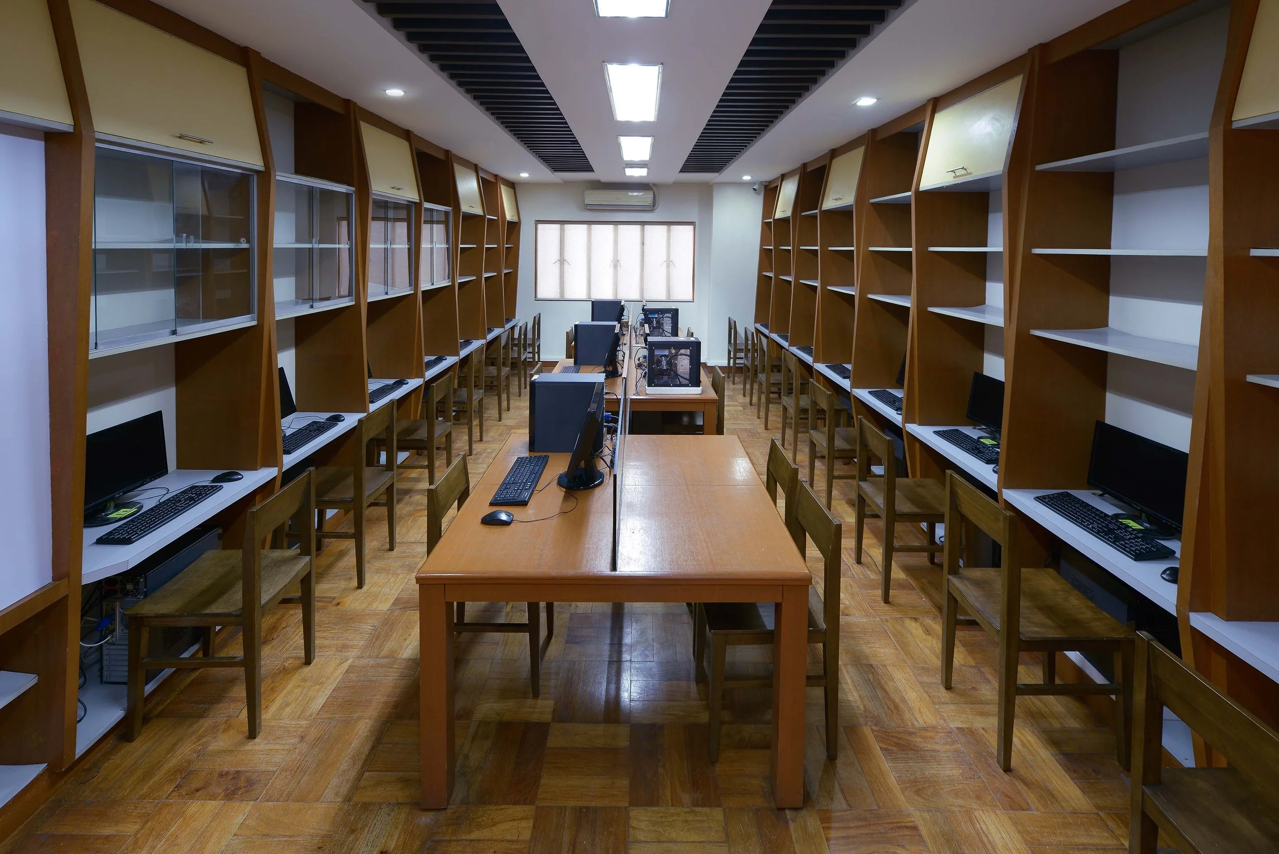 A computer lab with multiple workstations along both sides of the room, each equipped with a desktop computer, monitor, keyboard, and mouse. There is a central wooden table with televisions or monitors on top of it. The room has wooden flooring, a la
