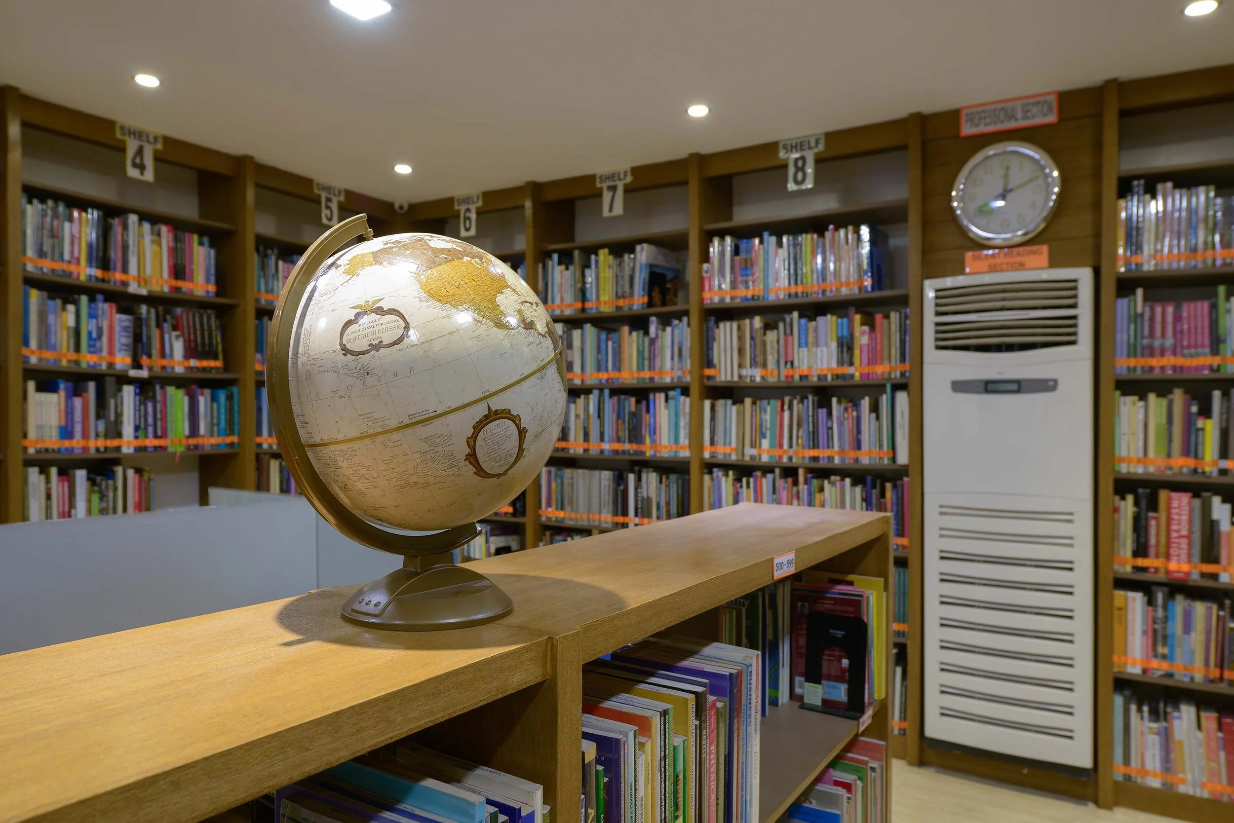 A globe on a wooden table in a library, with rows of bookshelves in the background labeled with shelf numbers, a clock, and an air conditioning unit.
