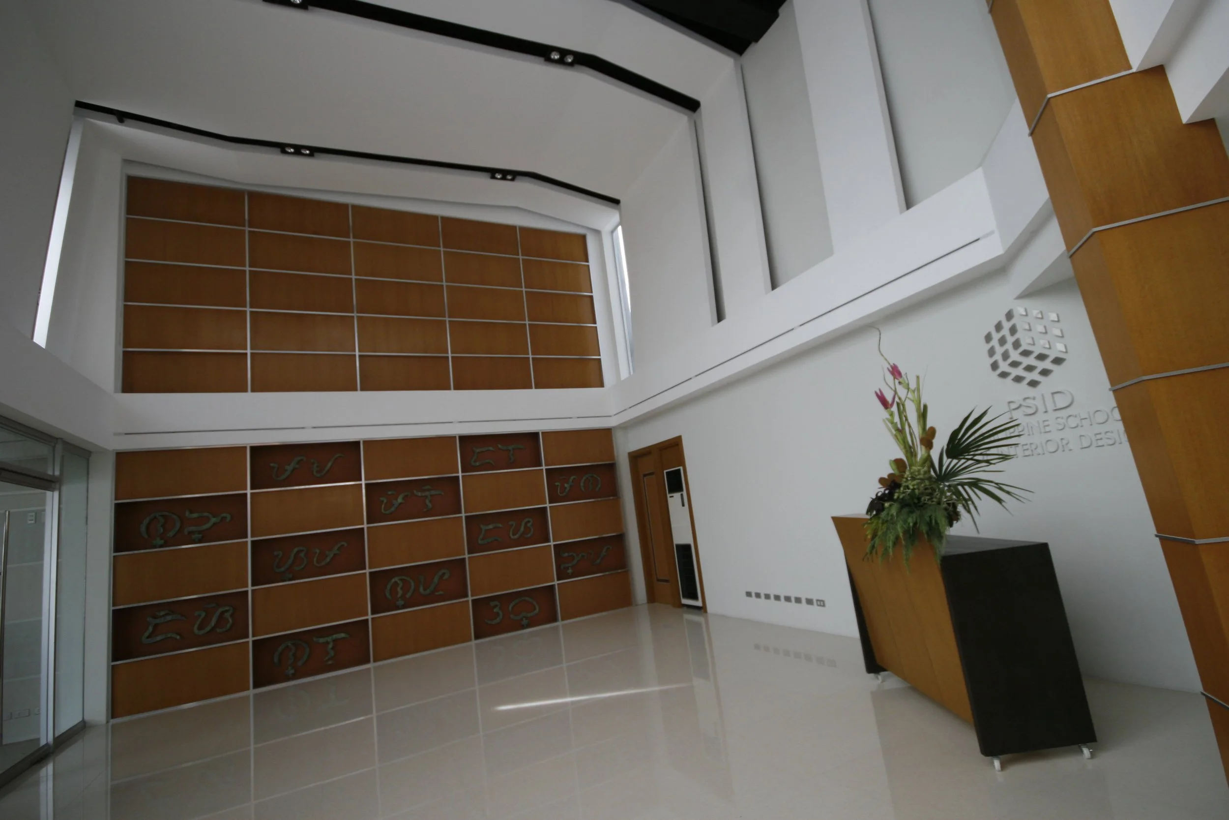 Modern interior space with a tall wooden reception desk, a bouquet of flowers, and a wall with wooden panels and metallic symbols. The ceiling has a large skylight with black support beams.
