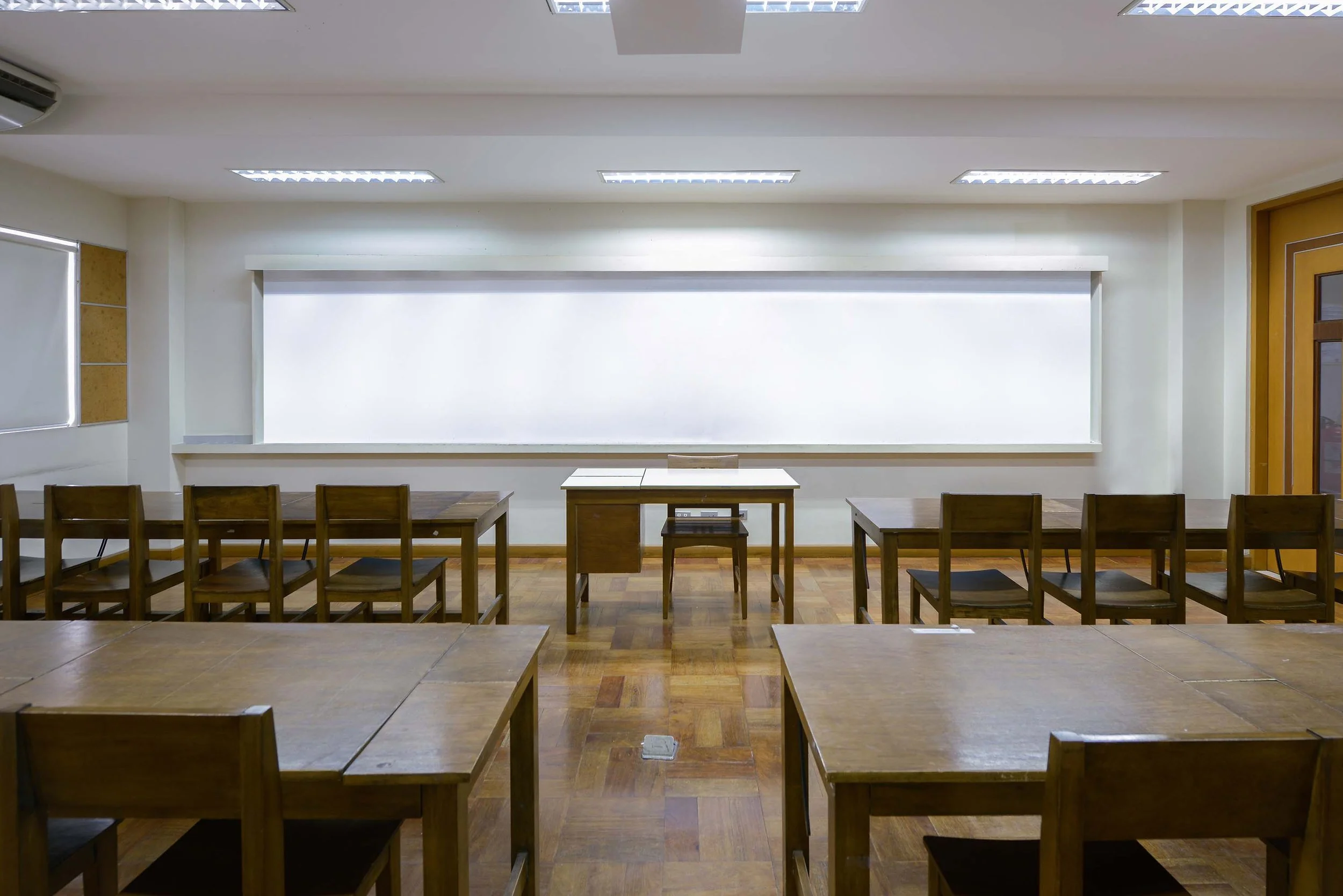 Empty classroom or conference room with wooden tables and chairs, whiteboard, windows, and ceiling lights.