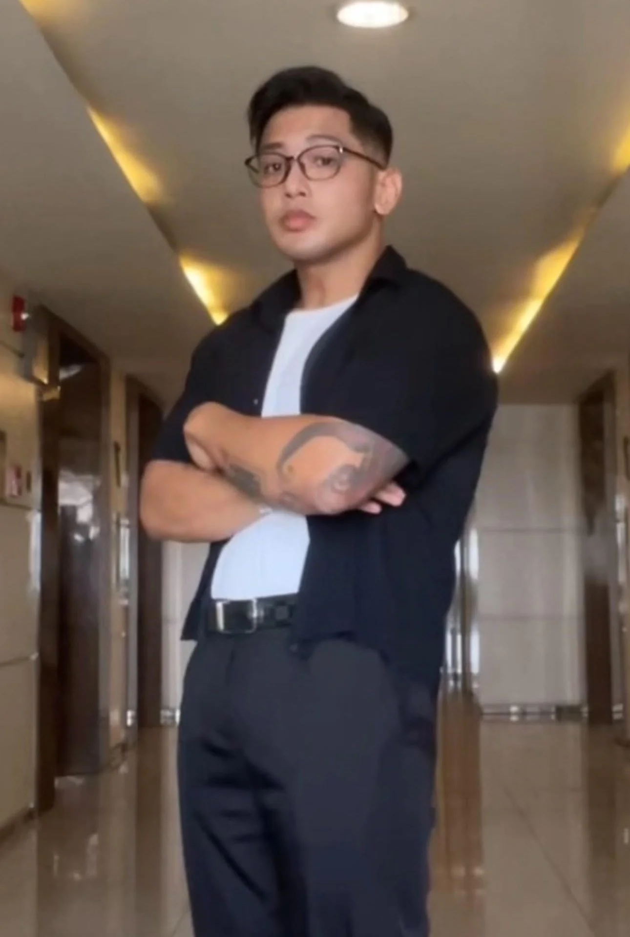 A young man with glasses and tattoos on his arms standing with crossed arms in a hallway with lockers.