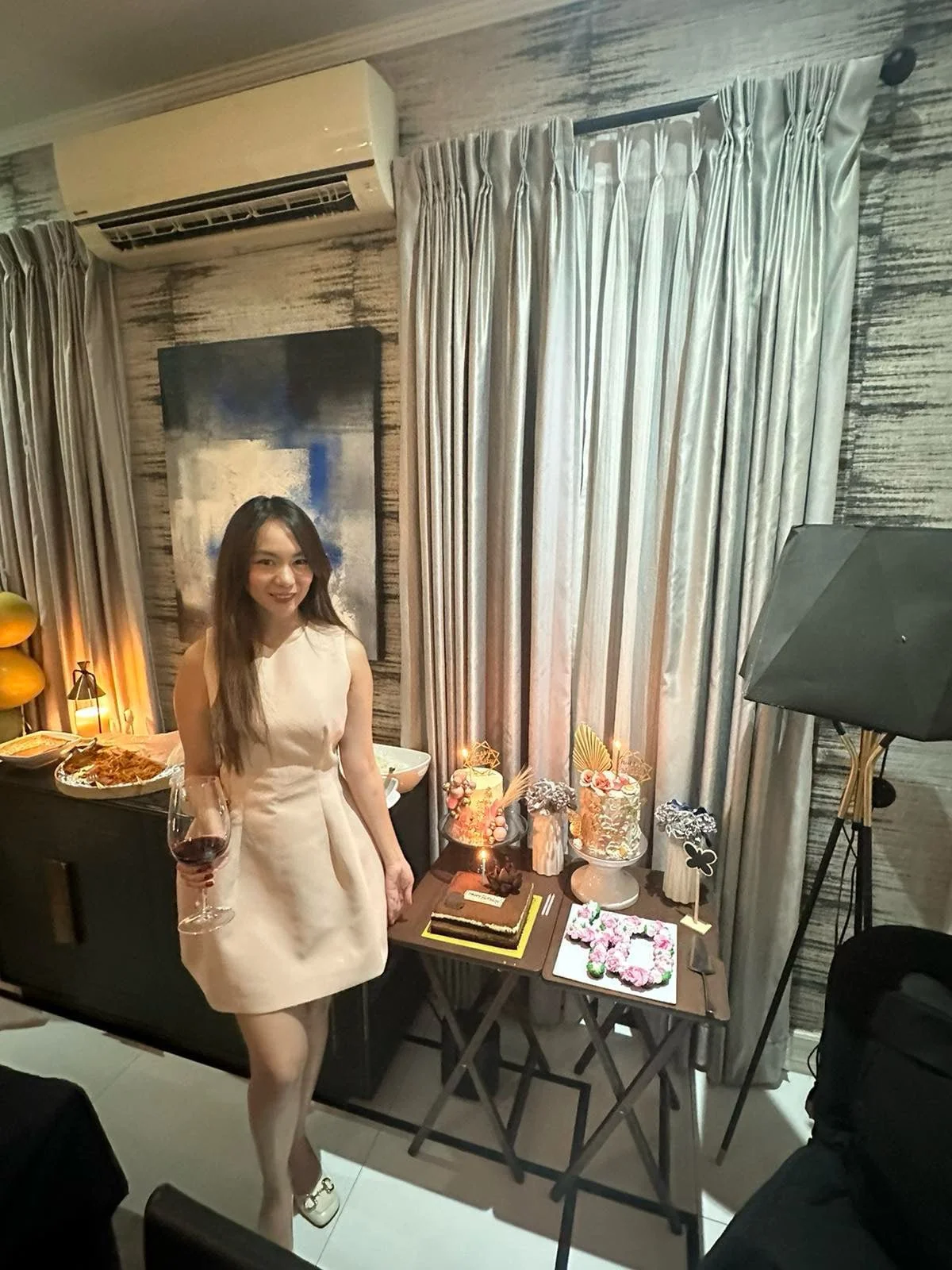 A woman in a white dress holding a glass of red wine is standing next to a table with cakes and desserts. The celebration appears to be a birthday or special occasion, with decorated cakes, flowers, and balloons in the background inside a room with curtains and textured walls.