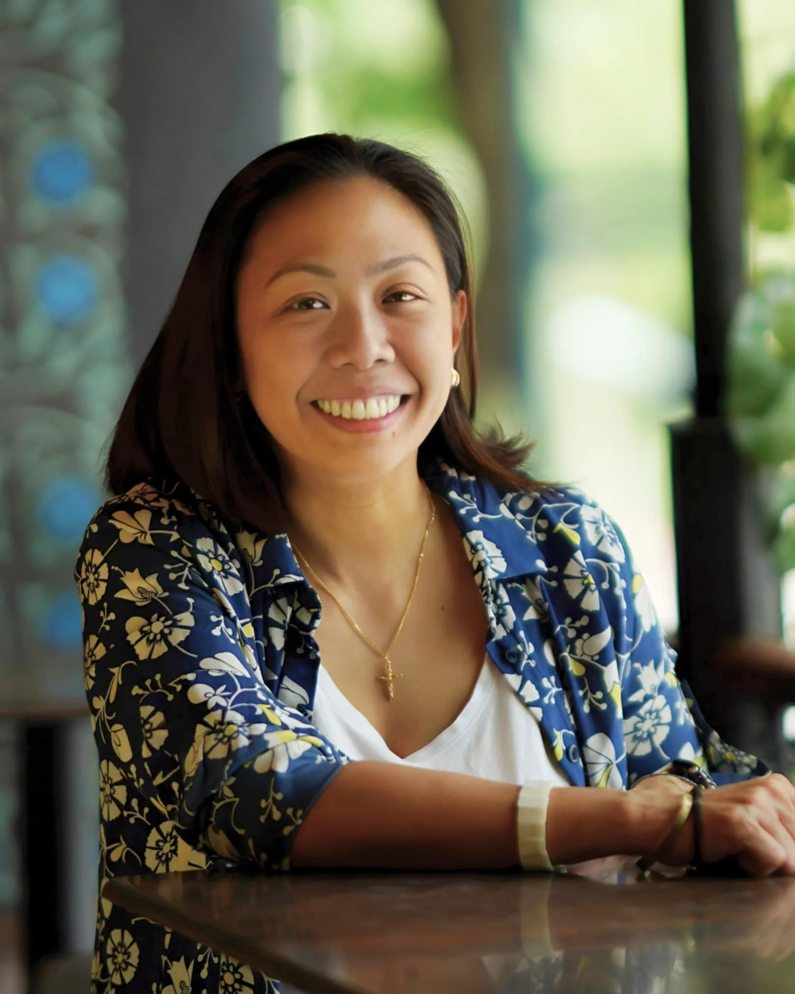 A young woman with dark hair smiling in a blue floral shirt and white top, sitting at a table in a bright, green outdoor setting.