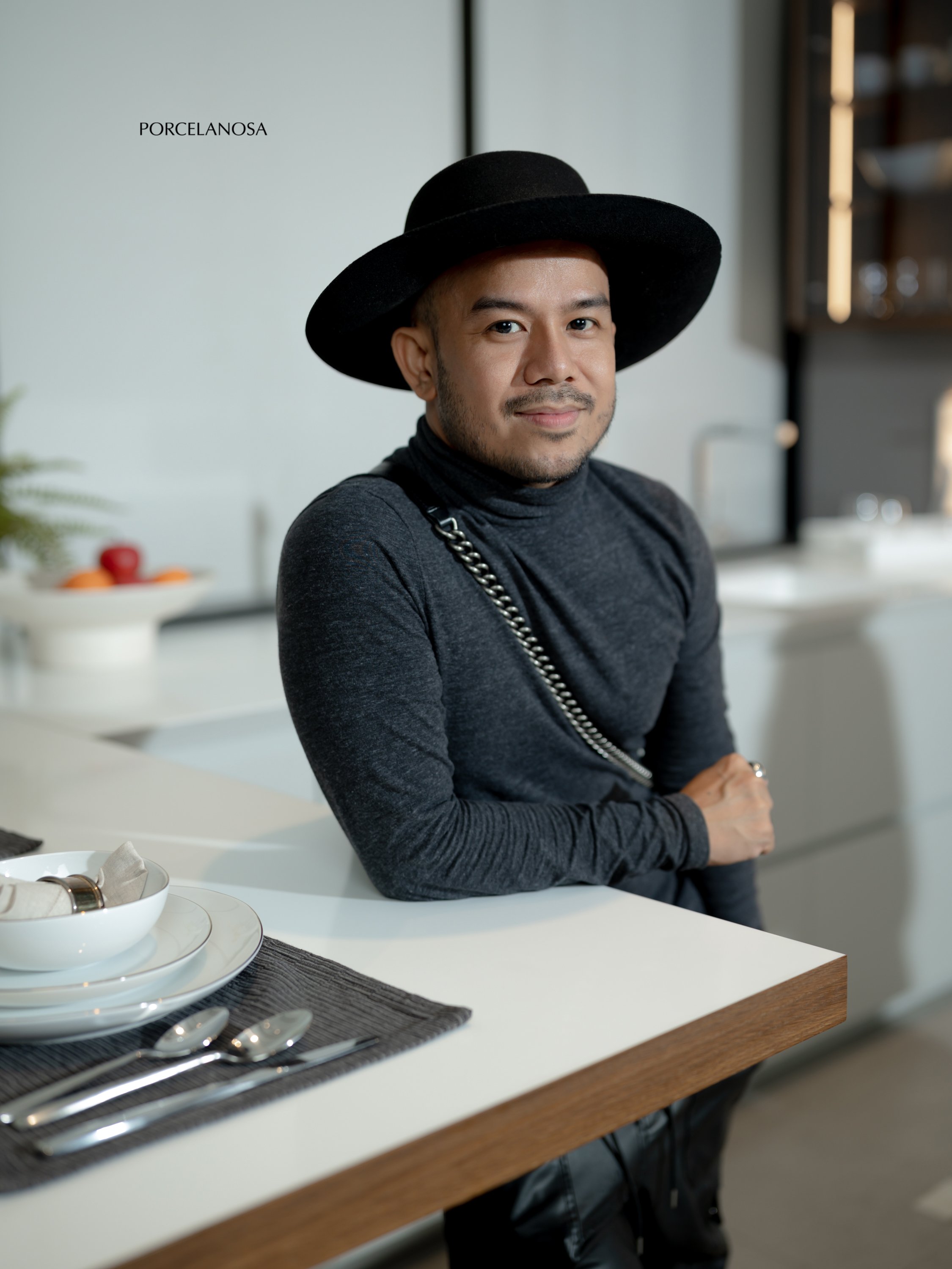 A man wearing a black hat and dark gray turtleneck standing in a modern kitchen, leaning on a white countertop with a bowl of utensils in front.