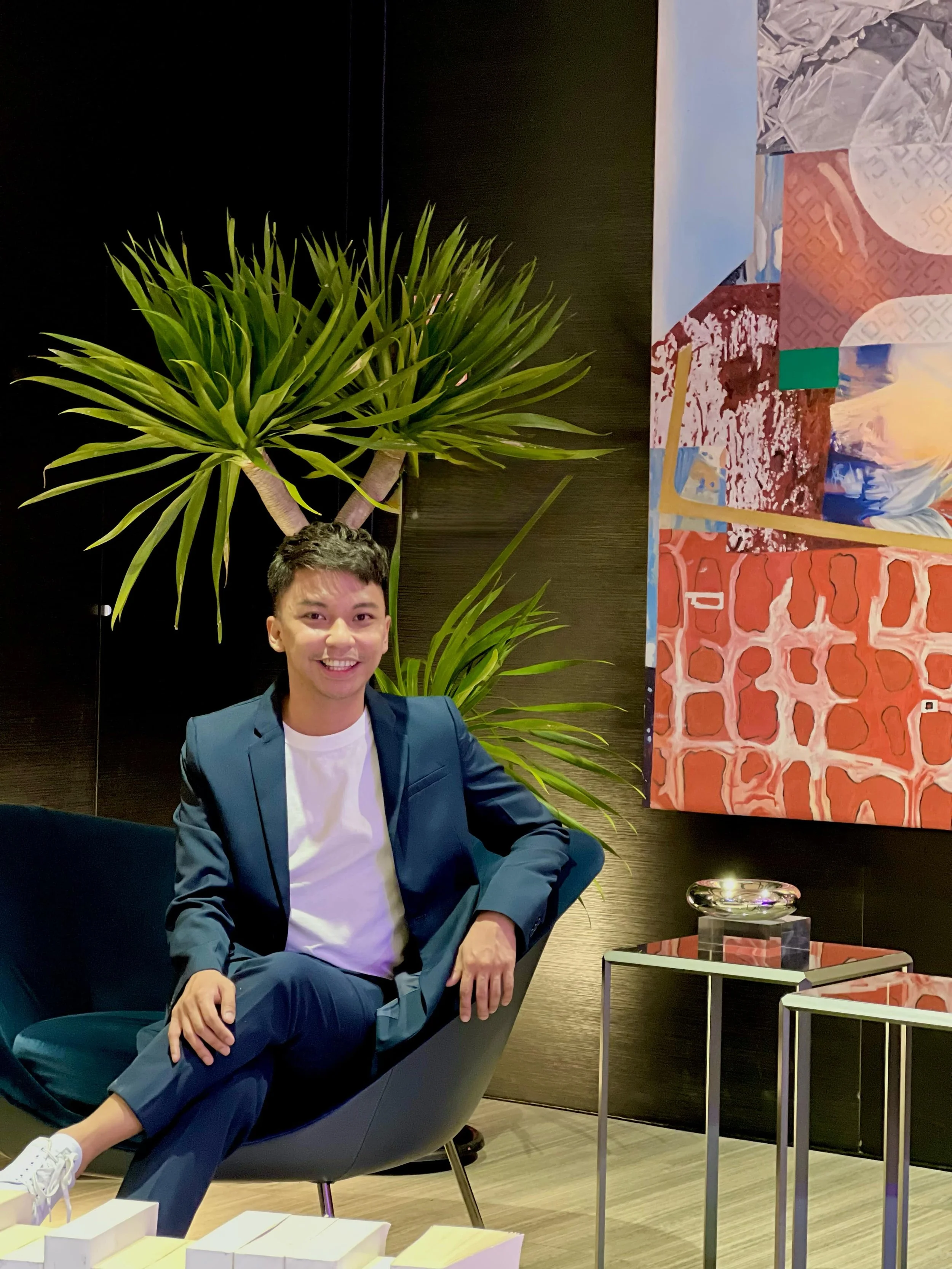 A young man in a blue suit and white t-shirt sitting comfortably in a modern black lounge chair, smiling, in an art-filled interior with a large colorful painting on the wall, green plants, and stylish furniture.