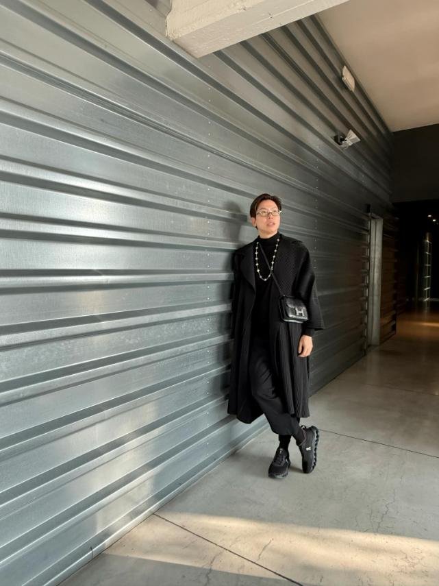 A person dressed in black standing against a metal roller shutter door in an indoor space, with a dark corridor visible in the background.