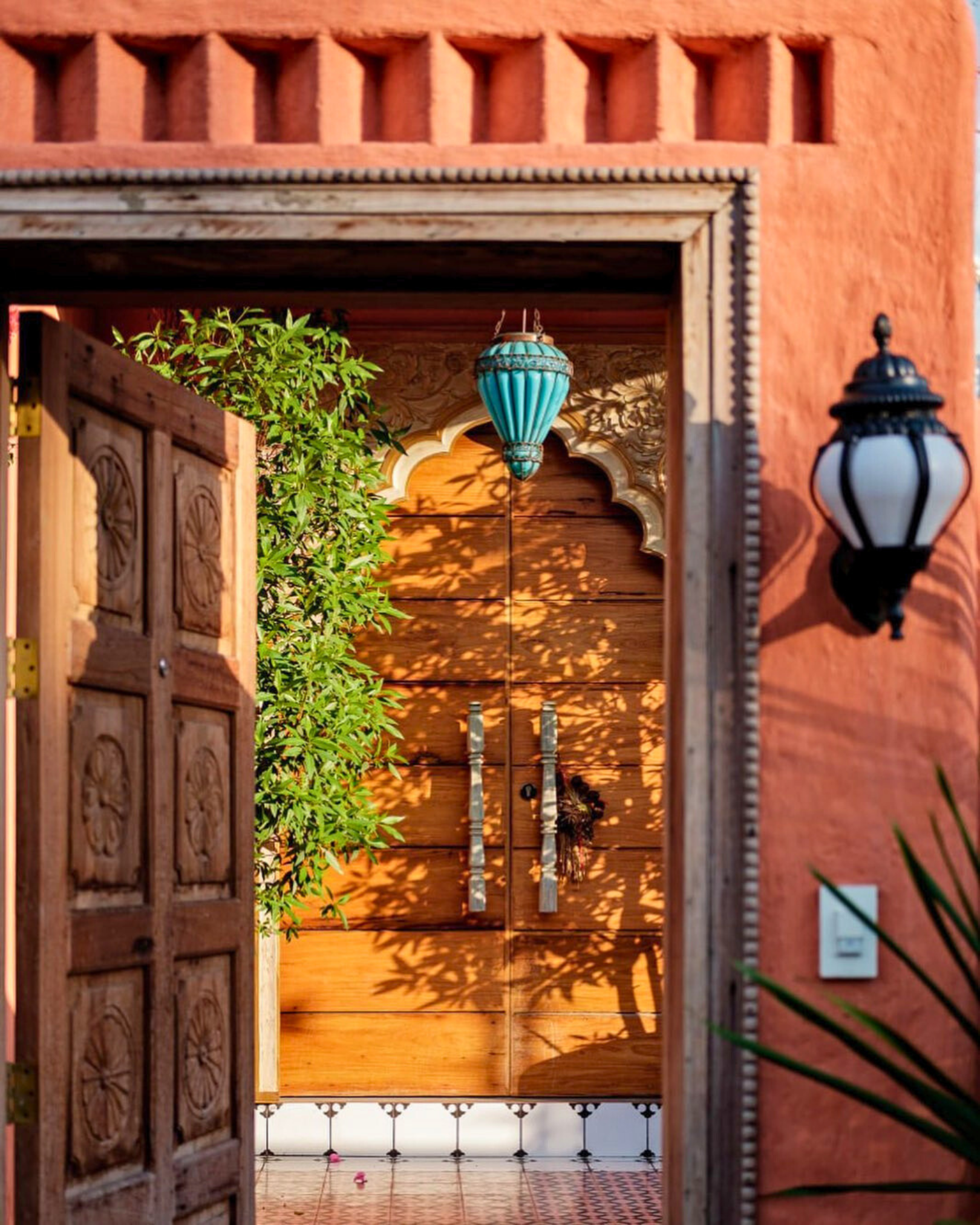 A decorative wooden entrance with an open door leading to a courtyard with tiled flooring, plants, and ornate hanging lanterns, including a blue lantern and black and white lanterns.