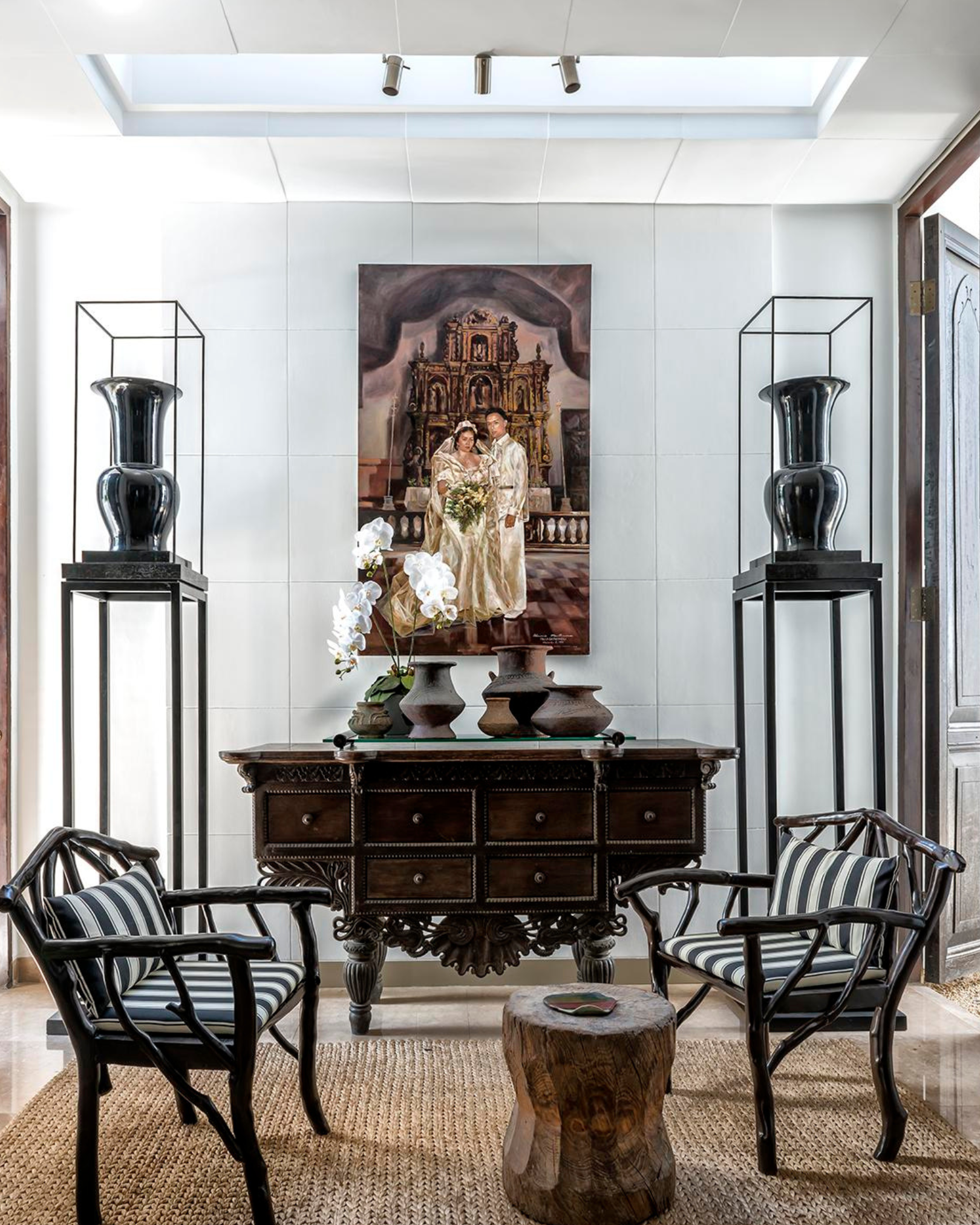 Interior of a room with a dark wood table in the center, four black chairs with striped cushions around it, a woven rug on the floor, a large framed painting of a couple and historical architecture on the back wall, two large black vases on black sta